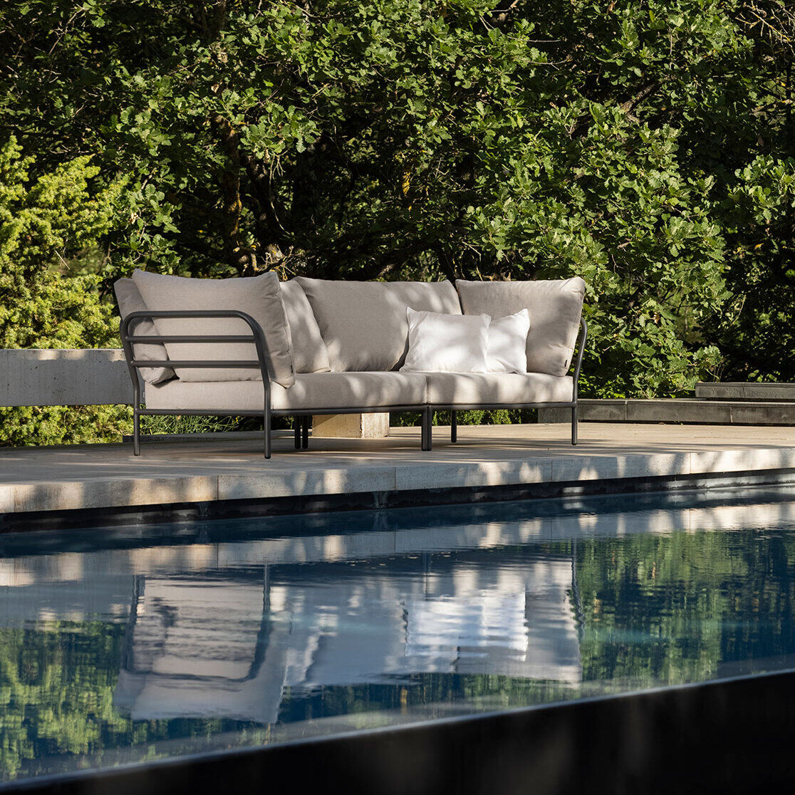 Modernes Outdoor-Ecksofa in einer Ecke am Pool, umgeben von grünen Bäumen und moderner Architektur.