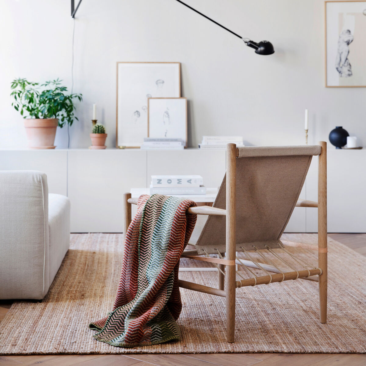 Ein stilvolles Wohnzimmer mit einer naturfarbenen Lammwolldecke 'Fri', die über einem minimalistischen Stuhl im skandinavischen Design liegt. Die neutrale Farbpalette des Raums enthält Beige, Braun, und Weiß Töne mit grünen Akzenten von Zimmerpflanzen. An der weißen Wand hängen dezente Kunstwerke und ein eleganter, schwarzer Wandstrahler ist sichtbar.