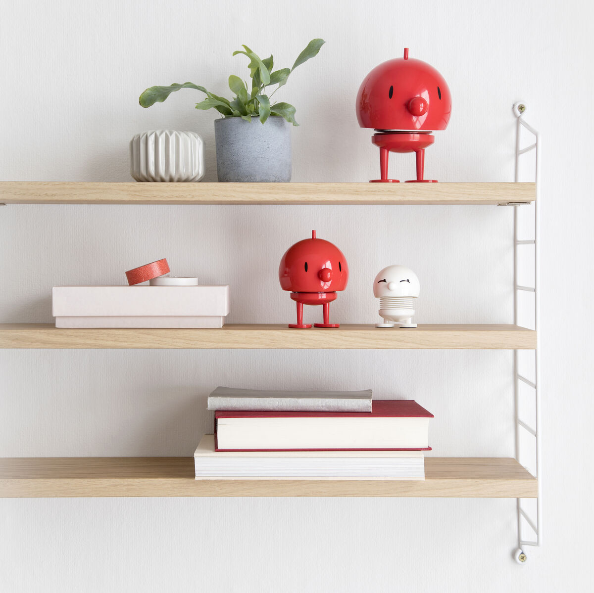 Auf einem minimalistischen Regal stehen zwei rote Hoptimist Bumble Figuren, umgeben von Büchern und einer kleinen Pflanze in einem grauen Topf.