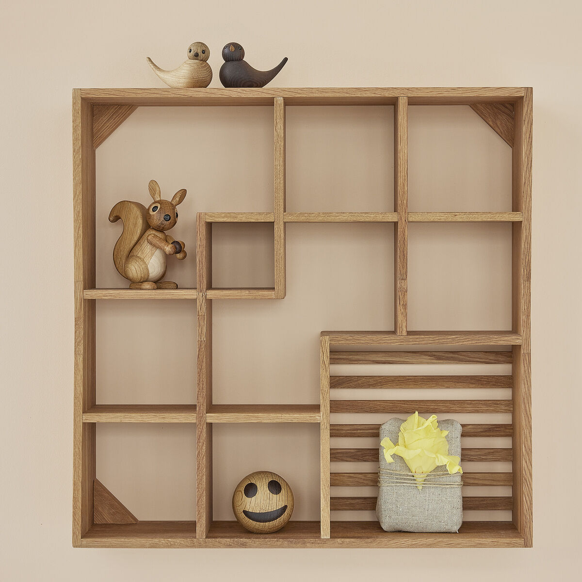 Holzfigur eines Eichhörnchens mit buschigem Schwanz steht in einem offenen Holzregal. Daneben befinden sich weitere kleine Holzfiguren und Dekorationen. Die Szene ist warm und freundlich gestaltet, ideal für Kinderzimmer oder herbstliche Dekoration.