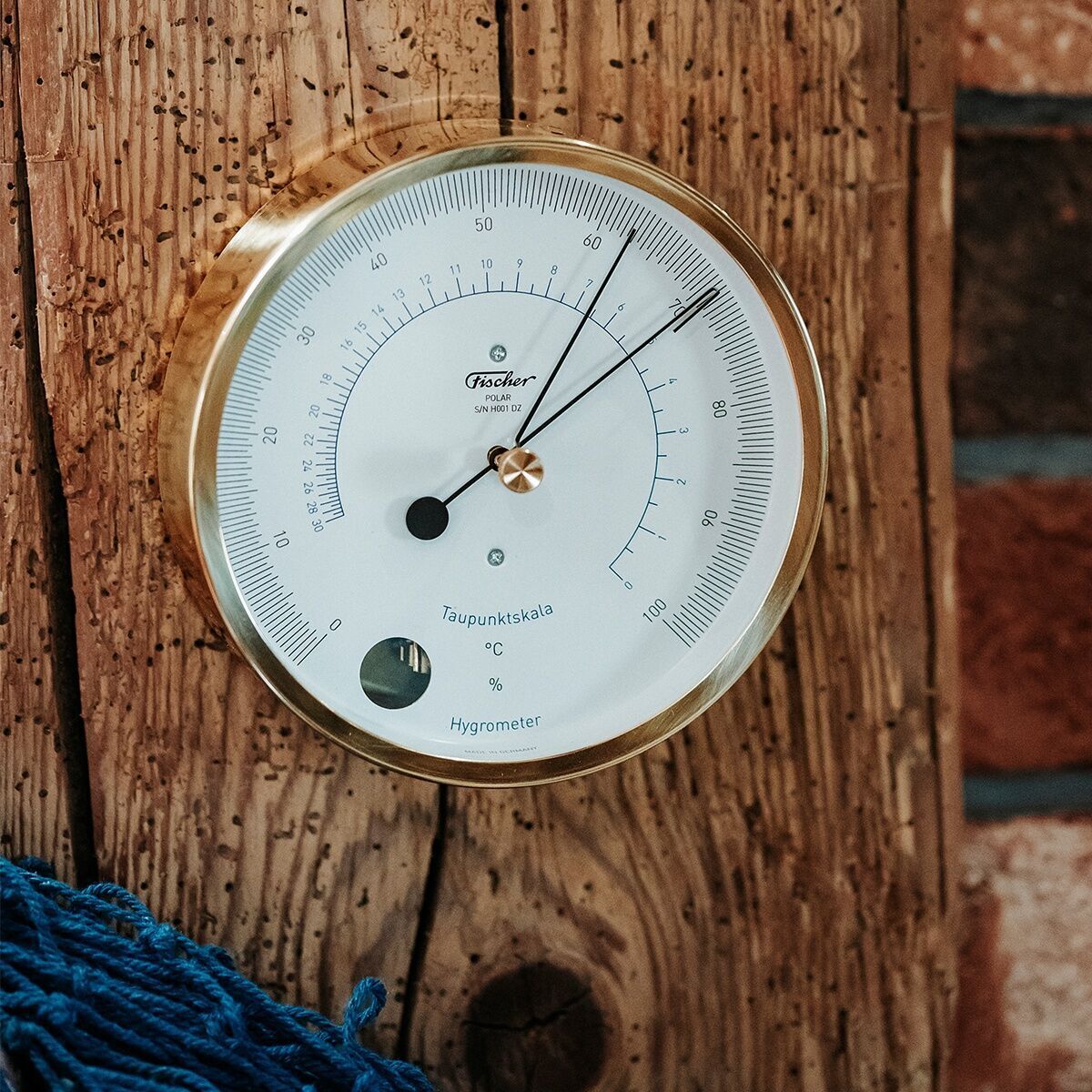 Das Bild zeigt ein ZEIT-Sonderedition POLAR Haar-Hygrometer mit messingfarbenem Rahmen, montiert auf eine Holzoberfläche. Darunter befindet sich etwas blaues Seil.