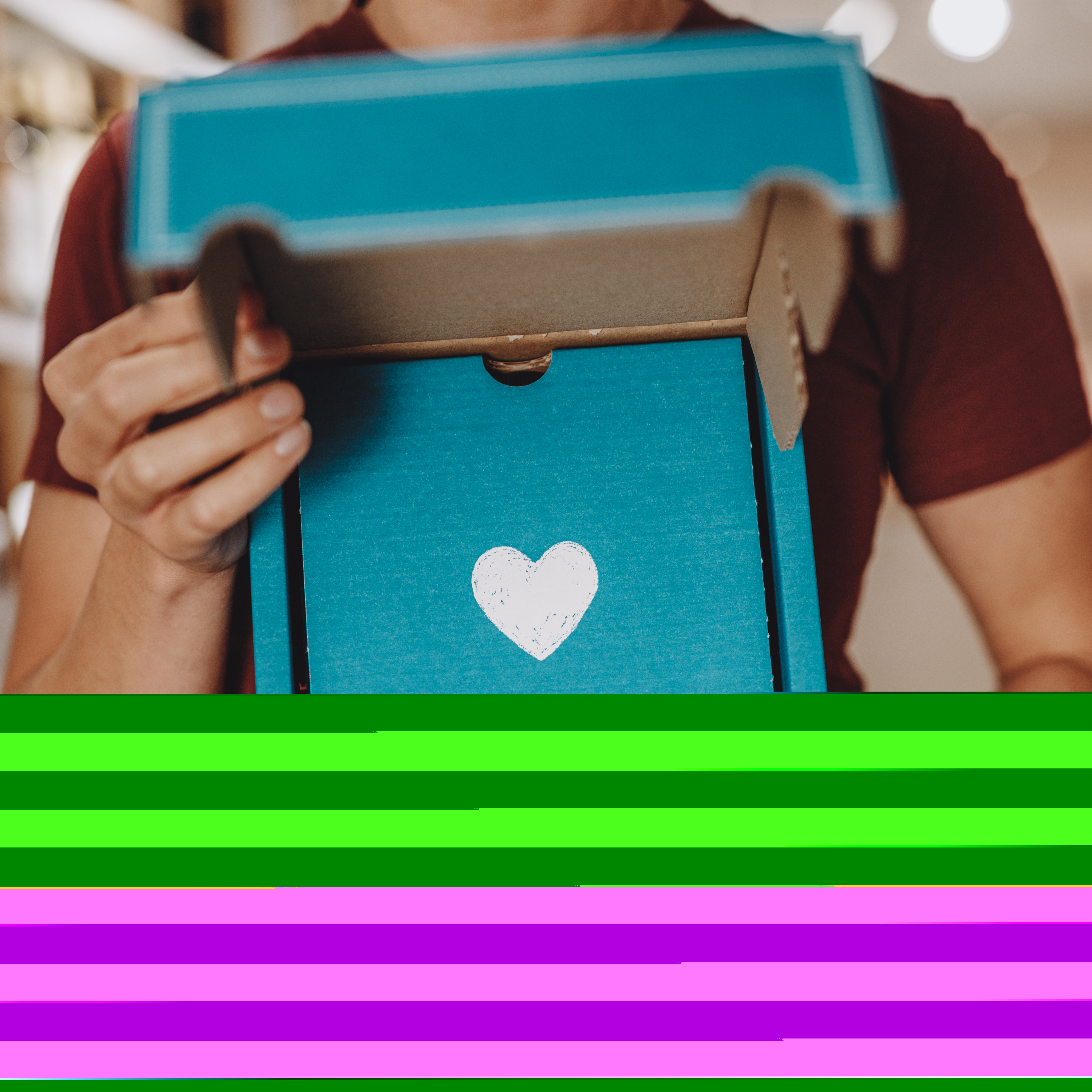 Eine Person hält ein türkisfarbenes Geschenkkarton-Set mit weißem Herzsymbol und der Aufschrift 'Mit Liebe handgemacht' in der Hand. Die Verpackung ist offen und wirkt einladend. Im Hintergrund ist ein Regal unscharf zu erkennen. Das Set enthält drei Flaschen mit je 250 ml Inhalt und eignet sich als Präsent für verschiedene Anlässe.