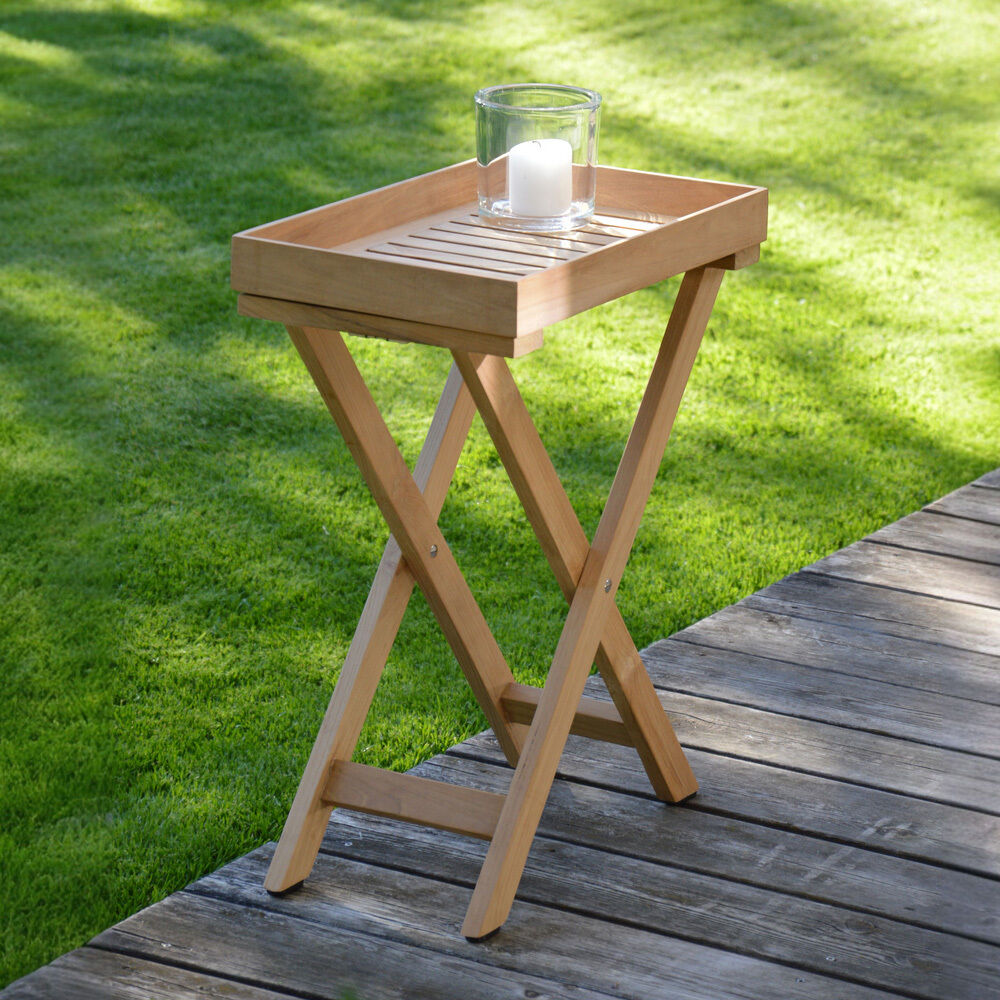 Der schmale, klappbare Beistelltisch »Chio« von Jan Kurtz aus recyceltem Teakholz steht auf einer Holzterrasse im Garten. Auf der rechteckigen Ablagefläche mit Tablettfunktion steht ein Windlicht mit weißer Kerze. Das Gras im Hintergrund ist sattgrün.