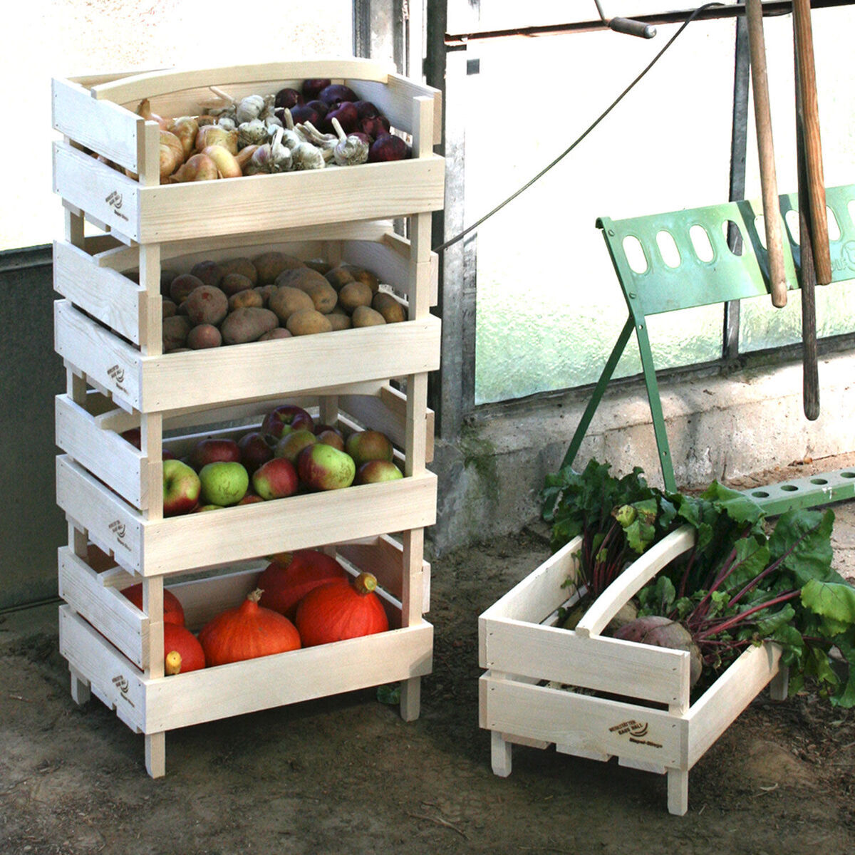 Mehrere gestapelte Holzkisten, gefüllt mit verschiedenen frischen Lebensmitteln wie Kartoffeln, Äpfeln, Kürbissen und Rüben, stehen vor einem Fenster. Eine grüne Gartenbank und Werkzeuge sind im Hintergrund zu sehen.