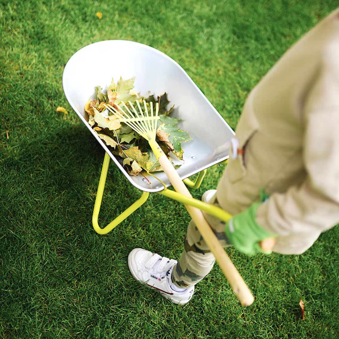 Detailreiche Szene im Garten: Ein Kind leert mit Gartenhandschuhen einen hellen Rechen und eine silberne Schubkarre, die mit Laub gefüllt ist. Die Schubkarre hat gelbe Griffe und Gestell. Das Setting vermittelt spielerisches Naturerlebnis und aktives Mithelfen beim Gartenarbeiten.