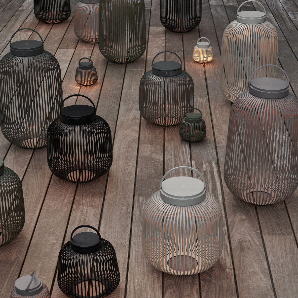 Mehrere blomus LITO LED-Leuchten stehen auf einer Holzterrasse. Sie variieren in Größe und Farbe – Granite Grey, Schwarz und Weiß. Die Leuchten erzeugen ein sanftes, dimmbares Licht. Ihr filigranes Lamellen-Design sorgt für elegante Schattenmuster und stilvolles Ambiente.