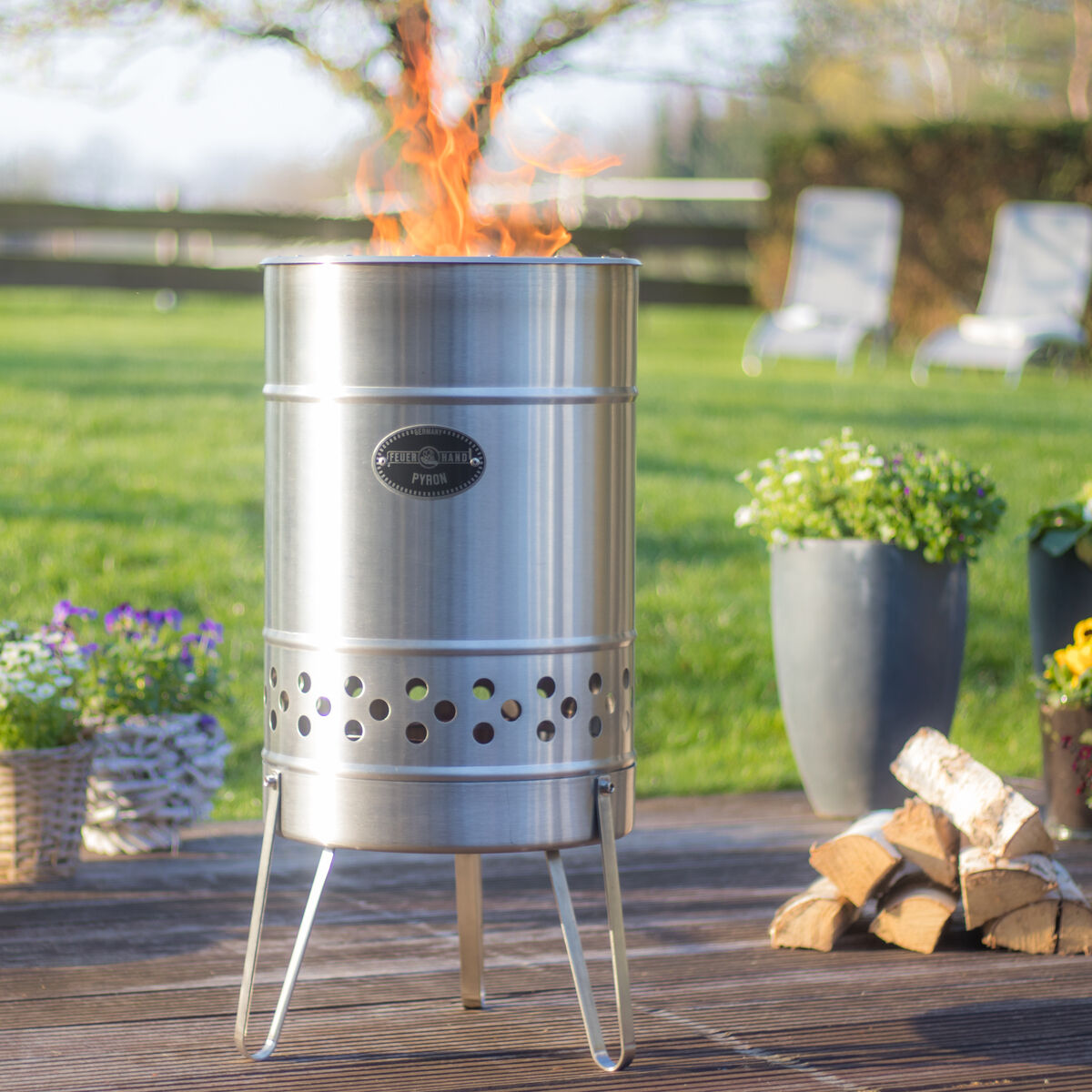 Feuertonne »Pyron« aus Edelstahl steht auf einer Terrasse, umgeben von Pflanzen und Holzscheiten. Im Hintergrund ist eine grüne Wiese mit Bäumen zu sehen. Die Feuertonne brennt und erzeugt Flammen.