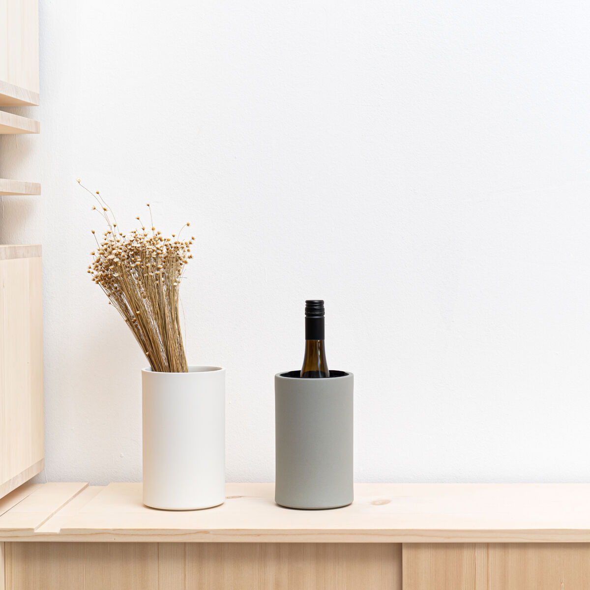 Zwei zylindrische Porzellanbehälter von Frau Caze/Carolin Zeyher stehen auf einem hellen Holzschrank vor weißer Wand. Einer ist betongrün und kühlt eine Weinflasche, der weiße dient als Vase mit Trockenblumen. Klare, minimalistische Gestaltung.