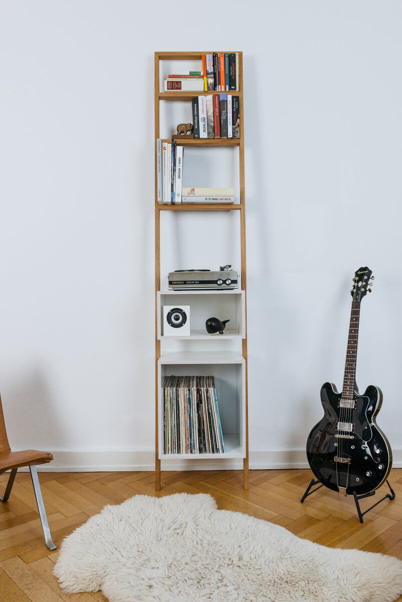 Ein schmales Leiterregal aus Holz mit weißem Korpus steht an einer weißen Wand. Das Regal enthält Bücher, Dekoartikel, eine Plattensammlung und ein kleines Lautsprechersystem. Daneben steht eine schwarze E-Gitarre. Auf dem Boden liegt ein weißer, flauschiger Teppich.