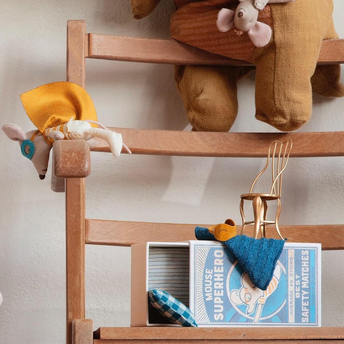 Eine kleine Maus als Superheld in einer Streichholzschachtel. Die Maus trägt ein Superheldenkostüm in Blau und Gelb. Neben der Schachtel steht eine Leiter aus Holz, an der eine weitere Stoffmaus hängt.