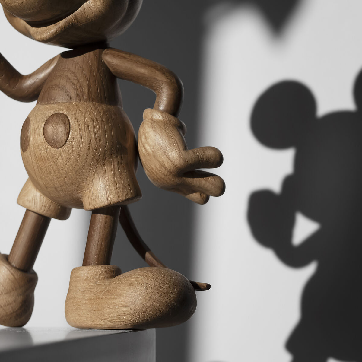 Links ist eine detailreiche Holzfigur von Mickey Maus zu sehen, gefertigt aus heller und dunkel gebeizter FSC-Eiche. Im rechten Bildteil fällt der markante Schatten der Figur auf eine weiße Wand, der die typische Silhouette von Mickey Maus zeigt. Hochwertiges skandinavisches Designstück.