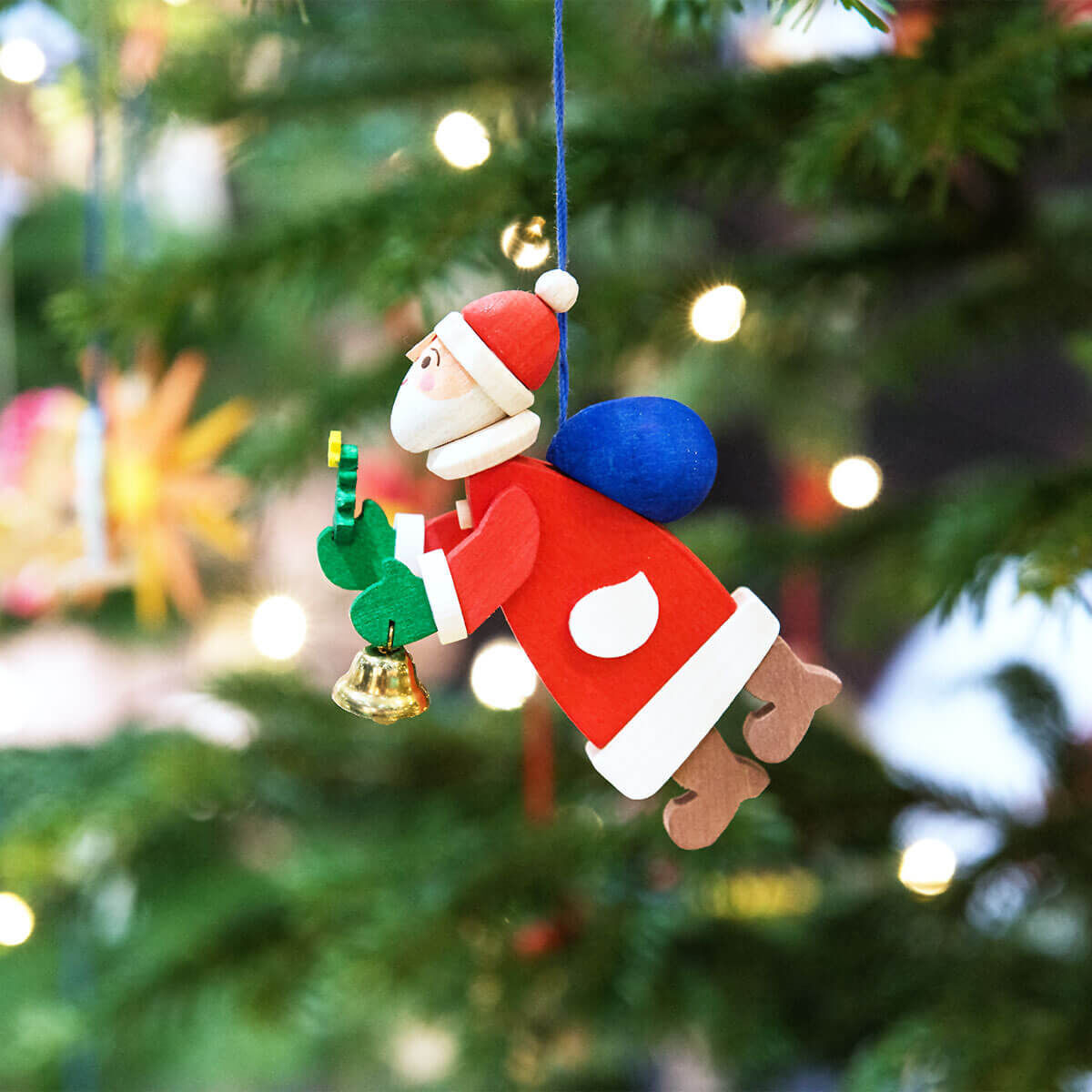 Weihnachtsbaumschmuck-Set mit drei handgefertigten Weihnachtsmann-Anhängern, die an einem Tannenbaum hängen. Die Weihnachtsmänner tragen rote Anzüge mit weißem Besatz, blauen Säcken und braunen Stiefeln. Der Baum ist mit Lichtern und anderen Dekorationen geschmückt.