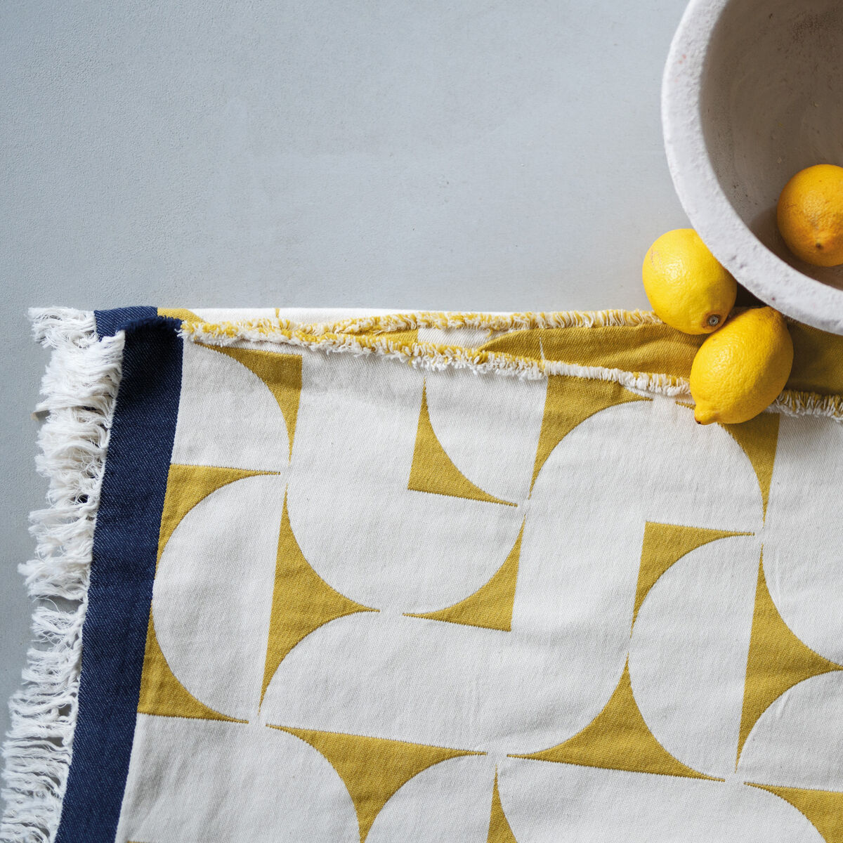 Baumwolldecke mit gelbem geometrischem Muster und blauen Fransen, dekoriert mit drei Zitronen, neben einer hellen Schüssel.