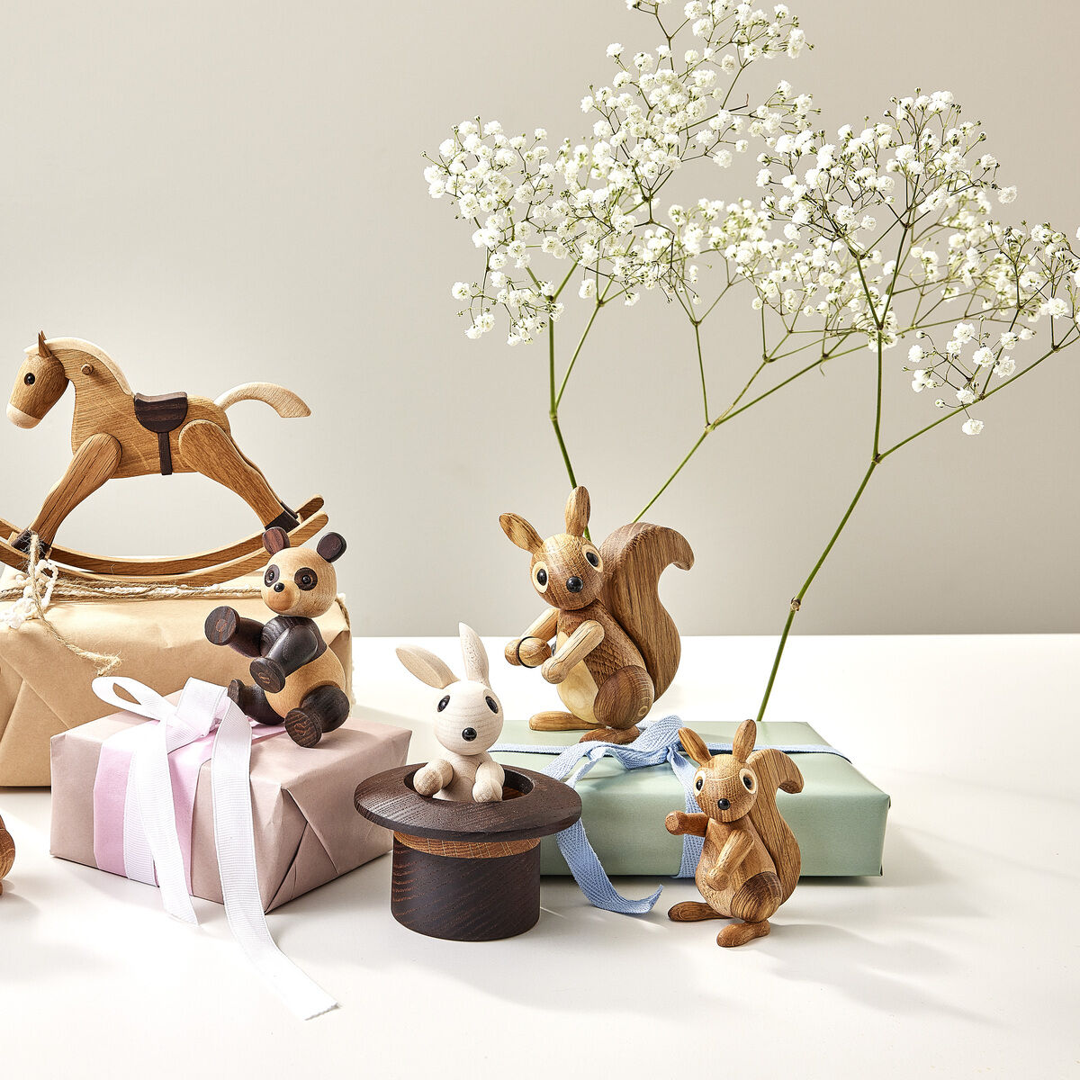 Mehrere liebevoll gestaltete Holzfiguren, darunter Eichhörnchen, Kaninchen, ein Pandabär und ein Schaukelpferd, stehen auf sowie neben in Pastellfarben eingepackten Geschenken auf einem hellen Tisch. Im Hintergrund befindet sich eine zarte Blume in einer schlichten Vase. Die Szene vermittelt eine freundliche, kindgerechte Atmosphäre mit natürlichen Materialien.