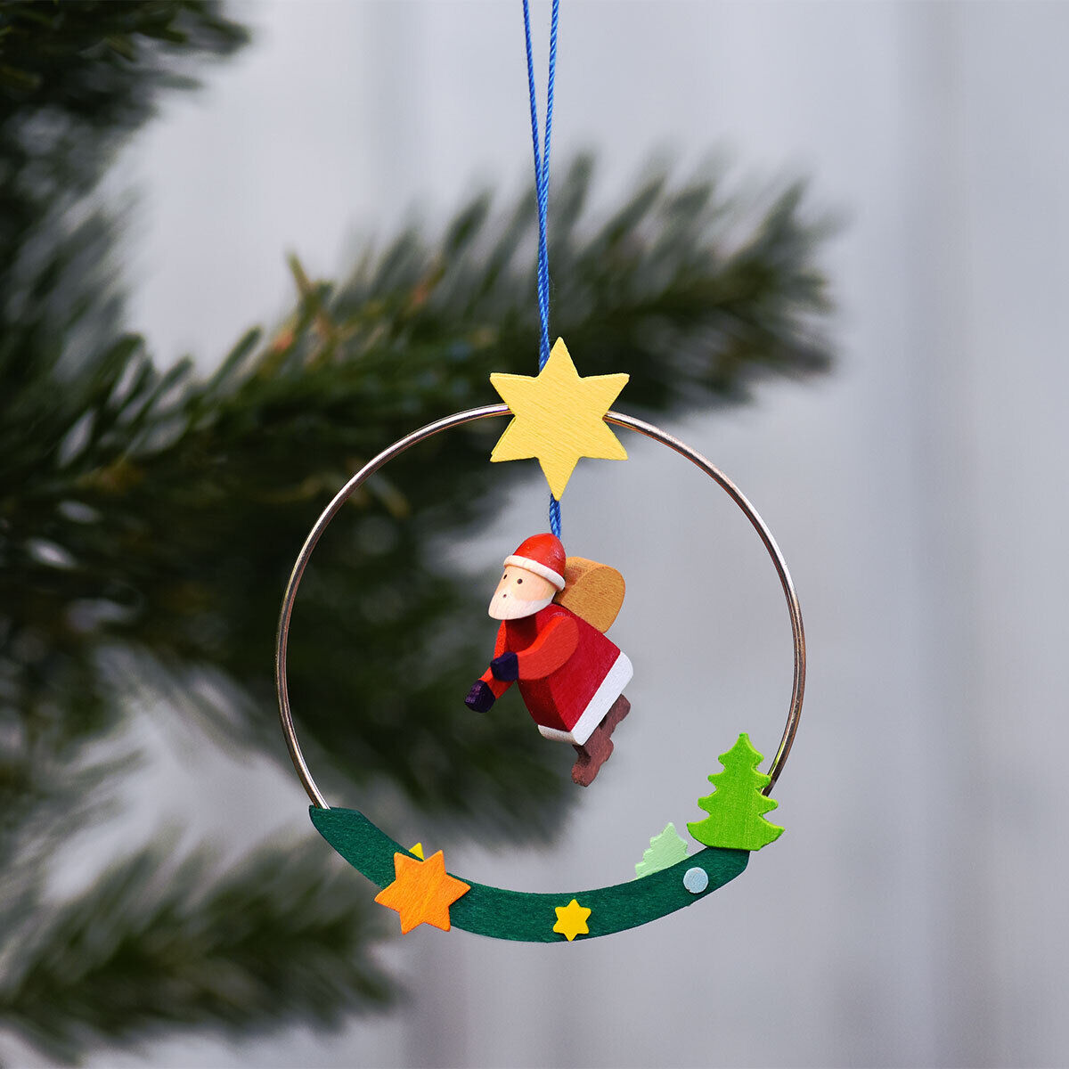 Ein handgemachter Weihnachtsanhänger in Form eines Weihnachtsmanns, der in einem Ring hängt. Der Ring ist mit Sternen und einem kleinen grünen Weihnachtsbaum verzierte. Der Anhänger ist überwiegend in den Farben Rot, Grün, Blau und Gelb gehalten. Hintergrund ist unscharfes Baumgrün und ein hellgrauer Hintergrund.