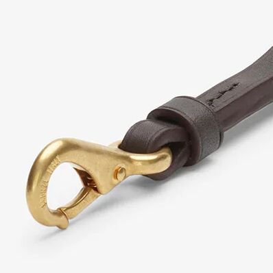 Detailaufnahme eines hochwertigen Schlüsselanhängers aus dunkelbraunem, pflanzlich gegerbtem Rindleder mit massiven goldfarbenen Messingbeschlägen. Der Karabinerhaken ist elegant geschwungen, die Lederkanten sind sauber poliert und fein genäht. Hintergrund schlicht, hellgrau.