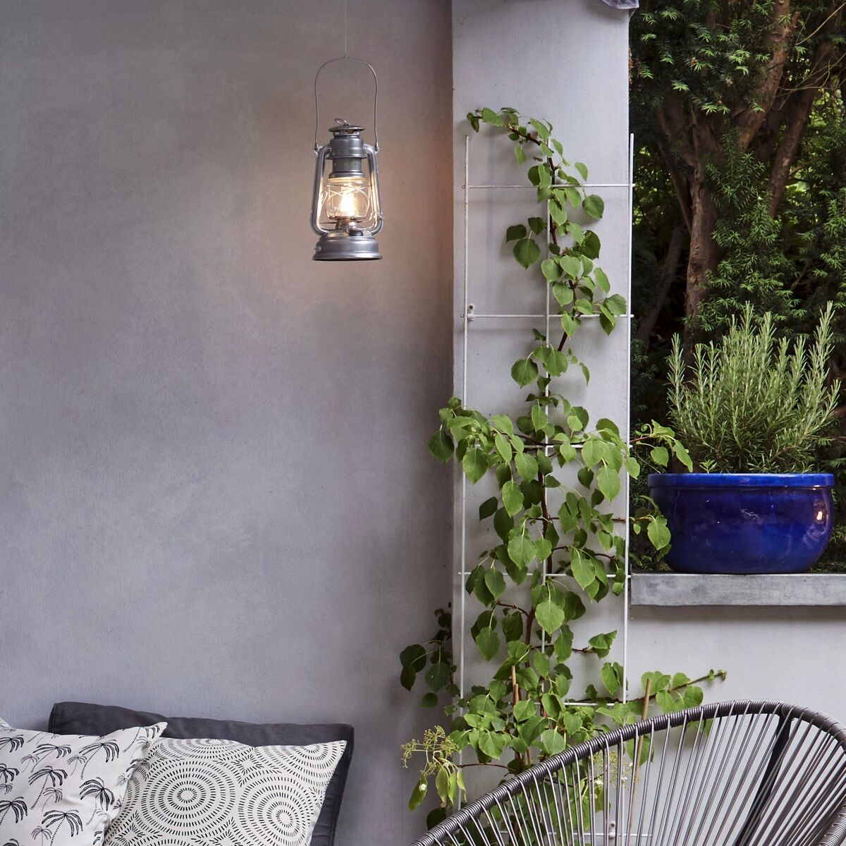 Eine moderne Terrasse mit gemütlicher Sitzecke. Eine LED-Laterne hängt an der Wand und eine weitere steht auf einem runden Hocker. Ein schwarzer, runder Rattansessel mit Kissen steht daneben. Kletterpflanzen wachsen an der Wand. Verschiedene Textilien und Pflanzen zieren den Raum. Der Boden ist aus Holz.