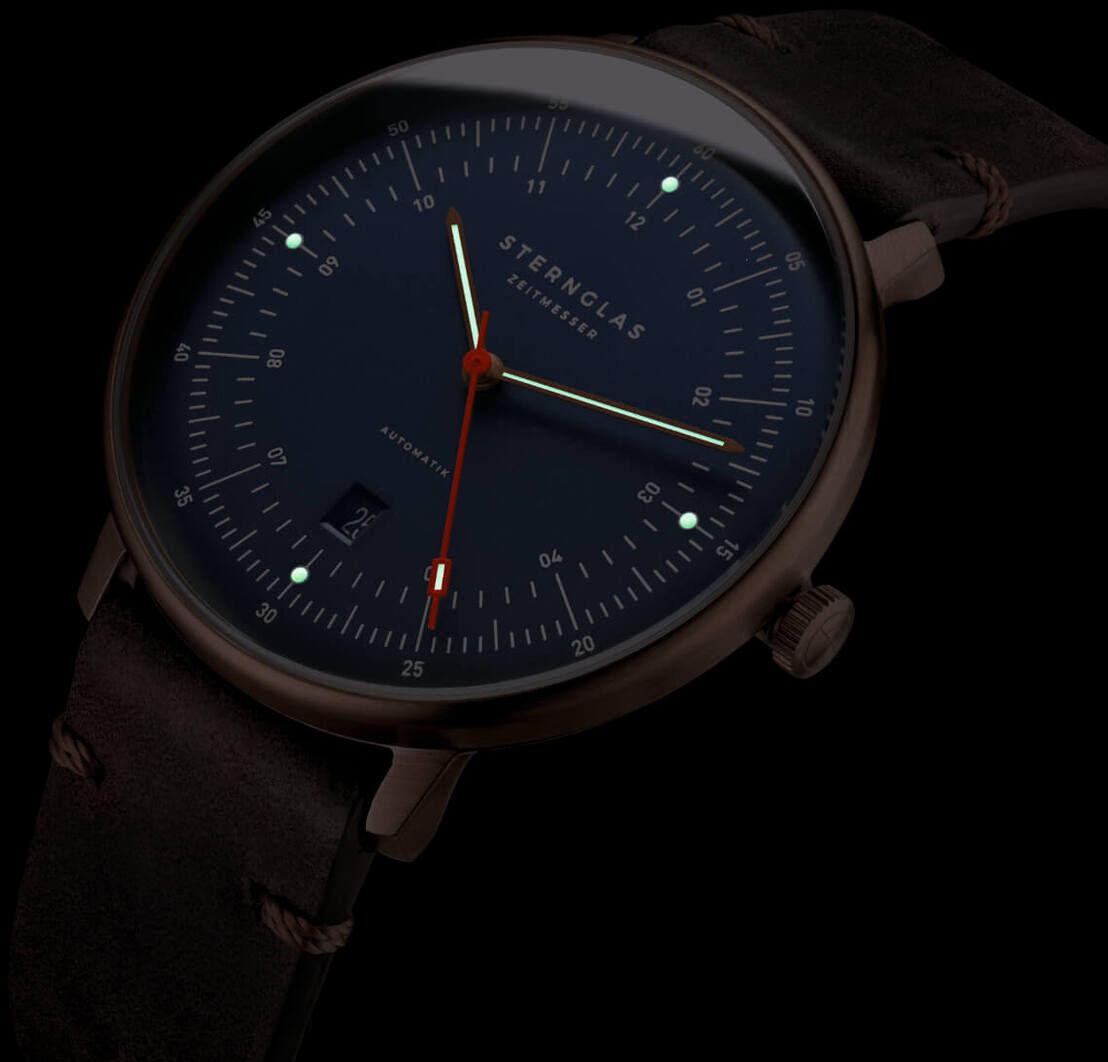 Eine ZEIT-Sonderedition Uhr »Hamburg« in Bronze mit einem dunklen Zifferblatt und einem Lederarmband. Das Zifferblatt ist schlicht gestaltet und zeigt klare, feine Markierungen. Zeiger in Rot und Weiß heben sich von der dunklen Oberfläche ab. Ideal für besondere Anlässe und als Geschenkidee.