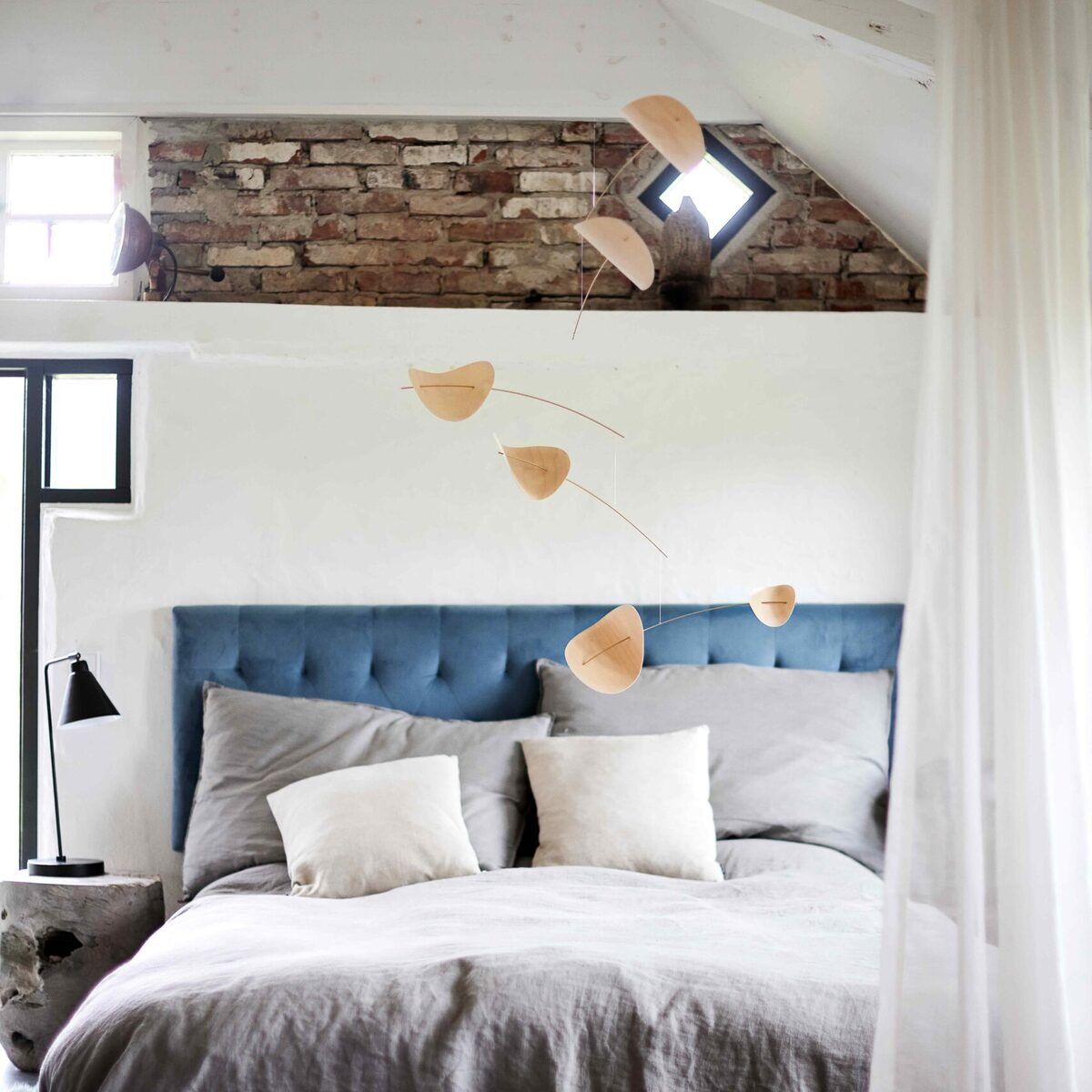 Ein minimalistisches Mobile mit dem Namen "Drifting Clouds", das aus Holz gefertigt ist und über einem Bett in einem modern dekorierten Schlafzimmer hängt. Die Bettwäsche ist in Grautönen gehalten, während das Bettkopfteil in Blau gestaltet ist. Ein gemauerter Ziegelfragment und Vorhänge in Weiß zieren den Raum.