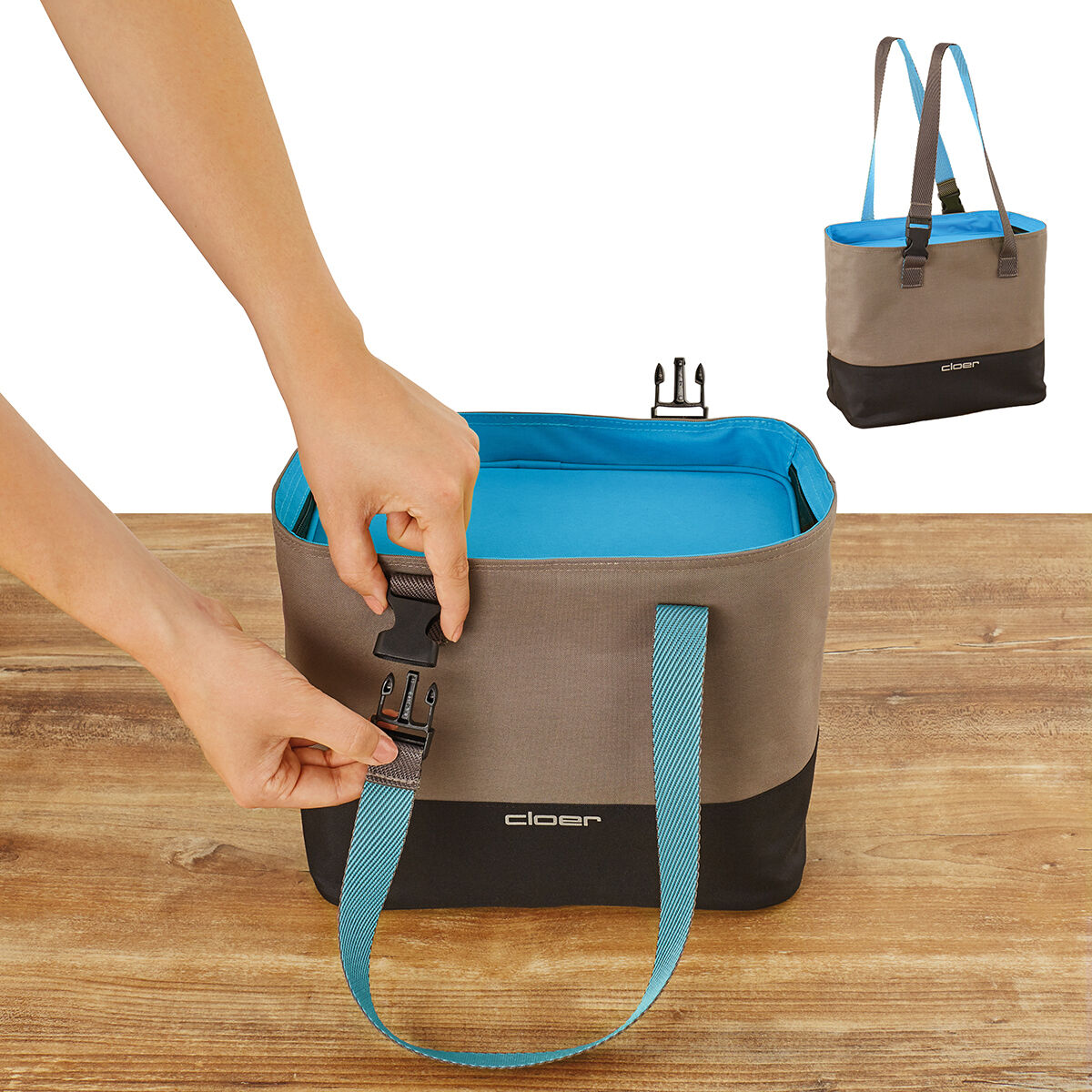 Eine Lunch-Tasche mit grauem und schwarzem Stoff und blauen Griffen wird auf einen Holztisch gestellt. Eine Hand ist dabei, den Verschluss der Tasche zu öffnen. Im Hintergrund ist eine kleinere Version der Tasche zu sehen.