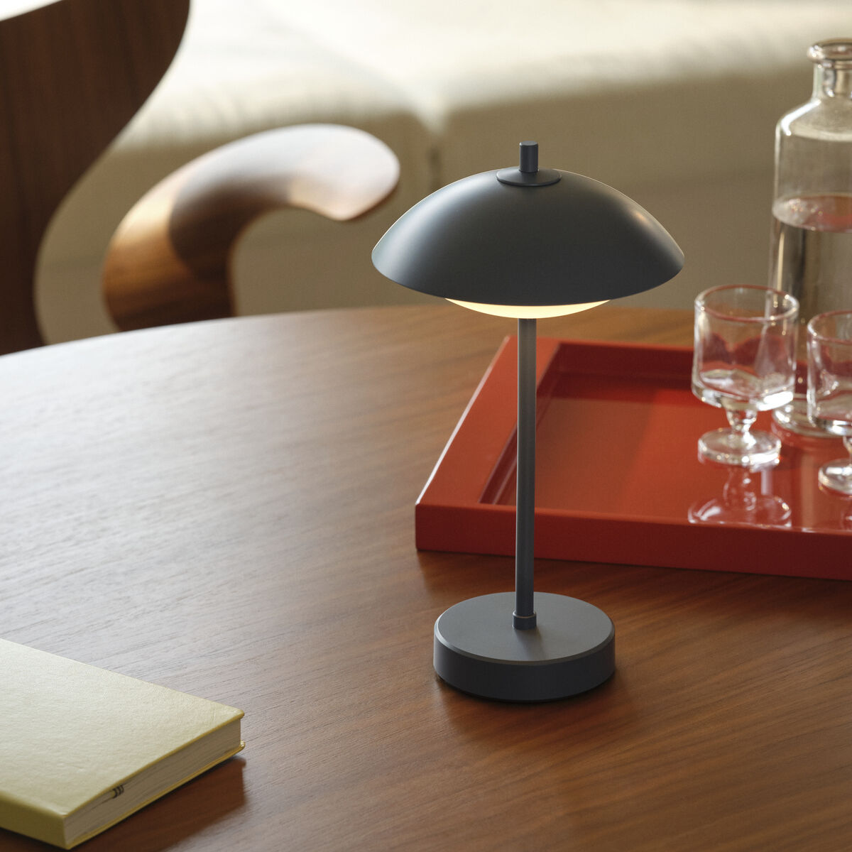 Auf einem runden Holztisch steht eine tragbare Lampe Clam™ von Fritz Hansen in mattem Blau. Daneben liegt ein gelbes Notizbuch. Im Hintergrund befindet sich ein rotes Tablett mit Gläsern und einer Glaskaraffe. Die Szene wirkt hell und stilvoll.