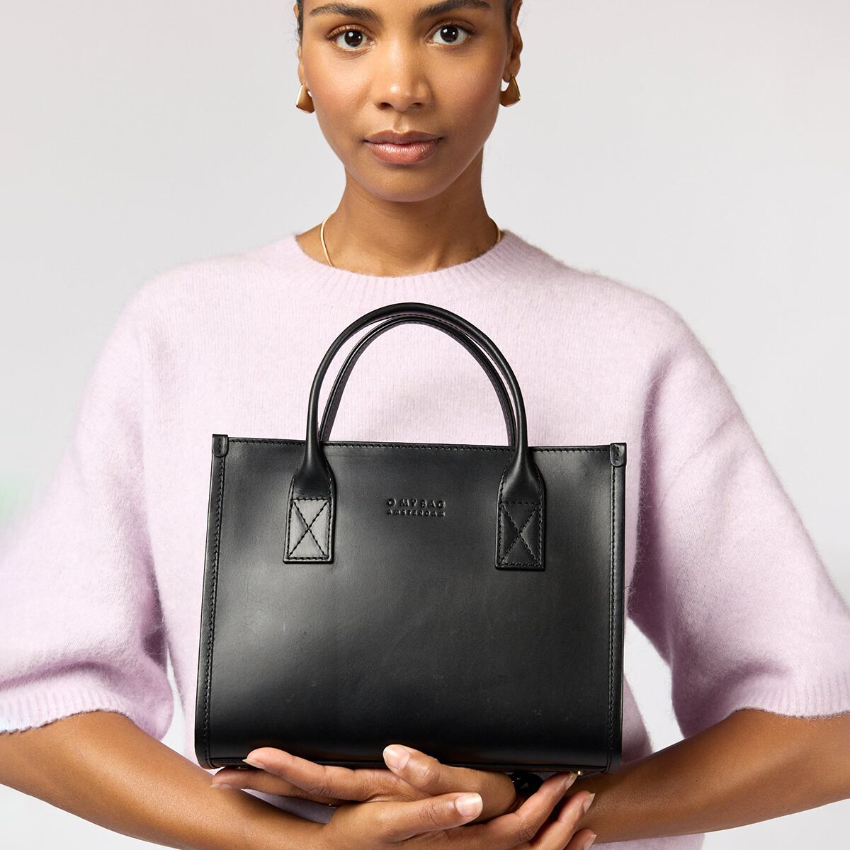 Eine Frau trägt eine schwarze, mittelgroße Handtasche aus Leder mit zwei Griffen vor dem Körper, während sie einen helllila Pullover trägt. Die Szene ist minimalistisch und fokussiert auf das stilvolle Design der Tasche »Jackie Midi«. Das Bild eignet sich zur Präsentation moderner Accessoires für Damen.
