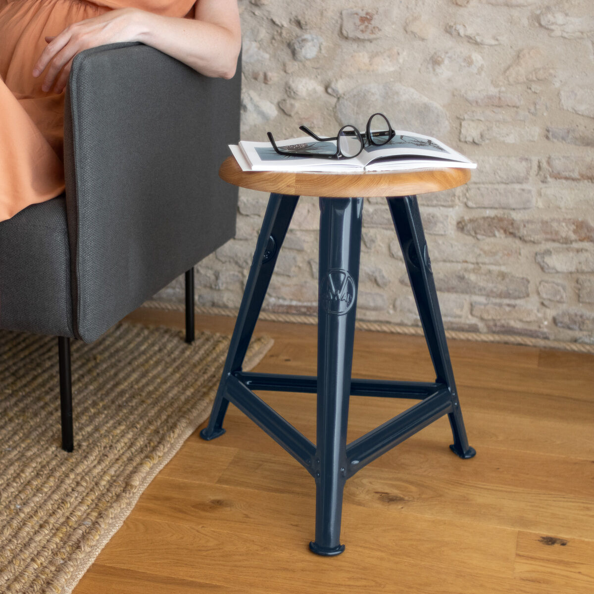 Ein handgefertigter blauer Stahlhocker der ZEIT-Sonderedition Rowac-Schemel in der Farbe »Blue Moon«, mit einer Holzsitzfläche, steht auf einem hellen Holzboden und neben einer grauen Couch. Auf dem Hocker liegen eine Brille und ein Notizbuch.