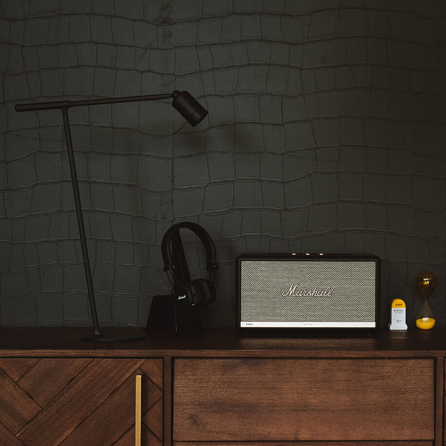Das Bild zeigt eine Tischlampe mit Halterung und weiteren Dekorelementen auf einem Holzschrank. Die Wand im Hintergrund wirkt wie mit Kunstleder verkleidet. Die Leuchte hat ein modernes Design und scheint ideal für ein stilvolles Interieur geeignet zu sein.