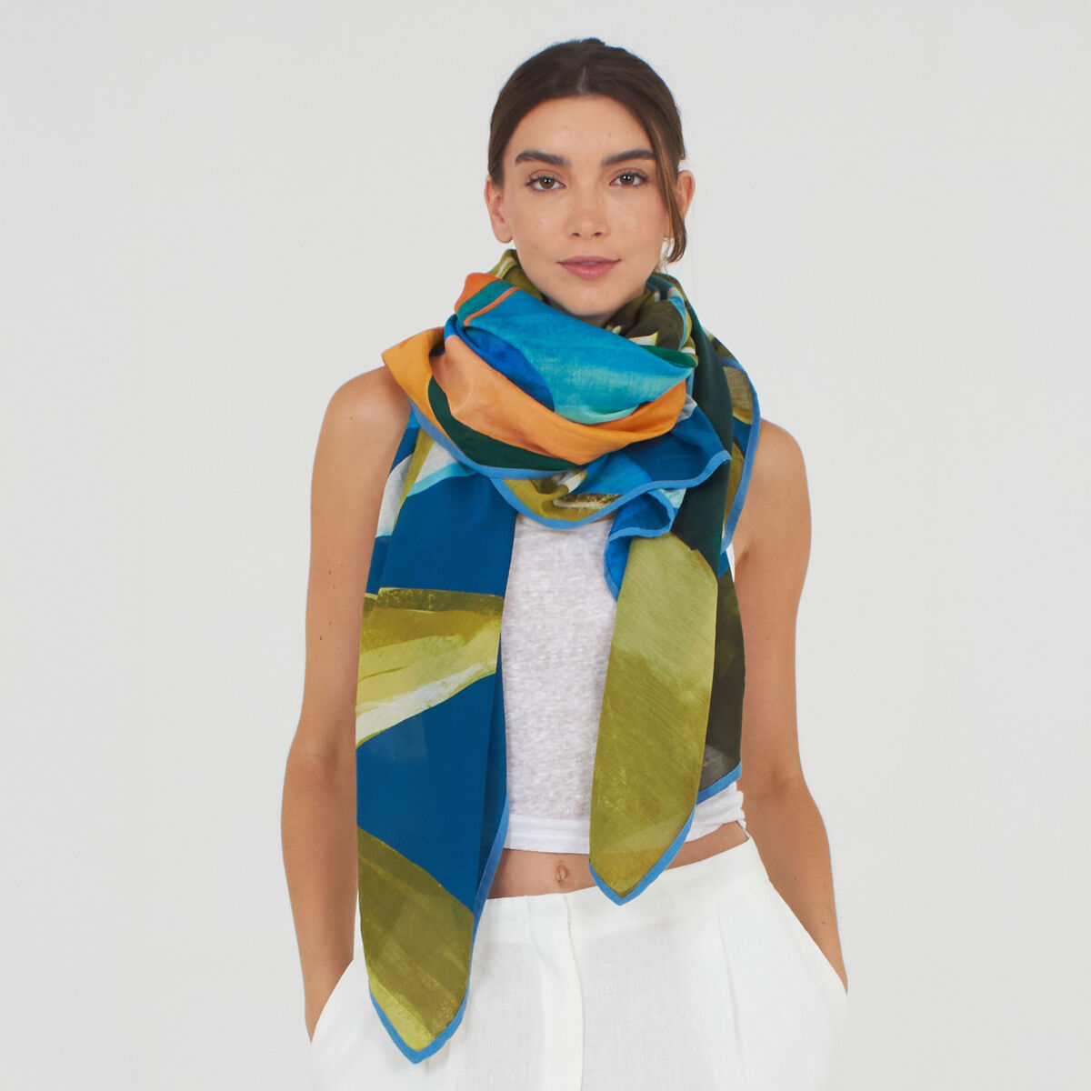 Frau trägt den Baumwoll-Seidenschal »Cassey« von Shalls Barcelona in Farbkombination Sea. Der Schal ist groß, mit abstraktem Muster in Blau-, Grün-, Gelb- und Orangetönen, und hochwertiger, leichter Stoff. Die Frau trägt ein weißes Top und weiße Hose.