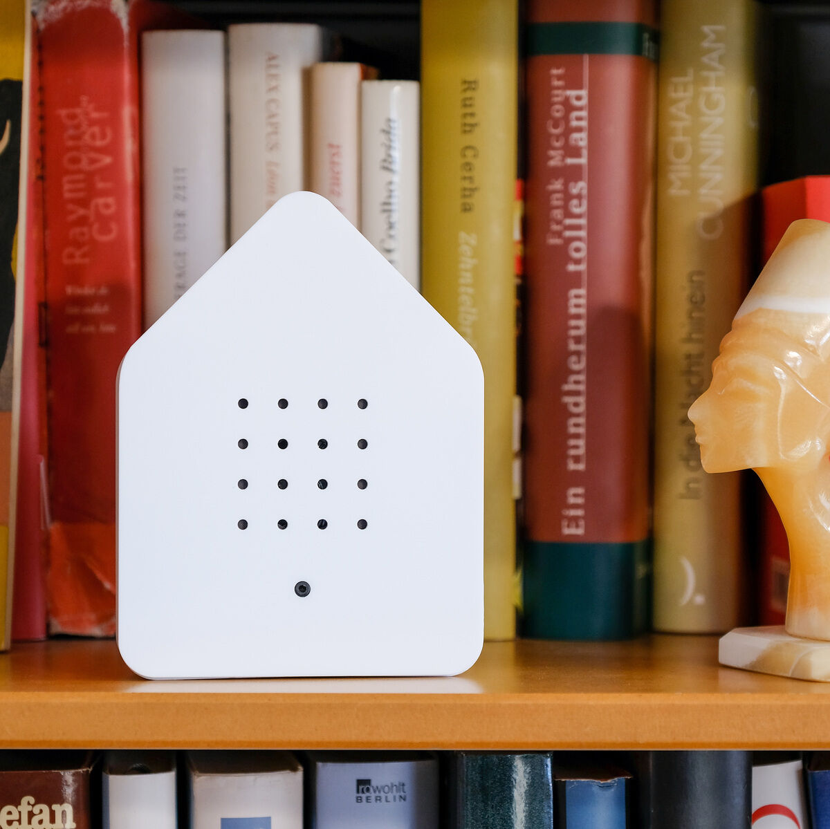 Ein stilvolles Design-Objekt namens 'Zwitscherbox' steht auf einem Bücherregal. Die Box ist weiß und hat die Form eines Hauses. Daneben steht eine gelbe Skulptur einer Büste. Im Hintergrund sind verschiedenfarbige Bücher zu sehen.