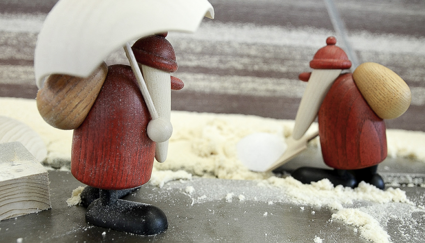 Zwei hölzerne Figuren mit roten Mänteln, schwarzen Schuhen und roten Mützen, eine hält einen weißen Regenschirm, beide tragen runde Rucksäcke, stehen auf einer mit Mehl bestreuten Fläche.
