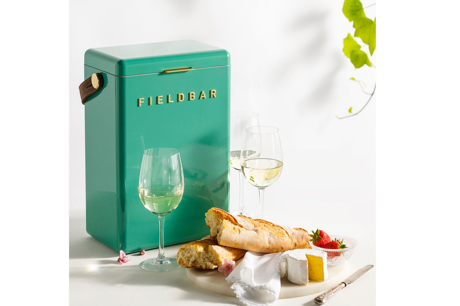 Eine grüne Drinks Box der Marke Feldbar steht neben einem gedeckten Tisch mit zwei Gläsern Weißwein, Baguette, Käse und Erdbeeren. Hintergrund mit Pflanzen. Ideal für Picknick oder Outdoor-Veranstaltungen.