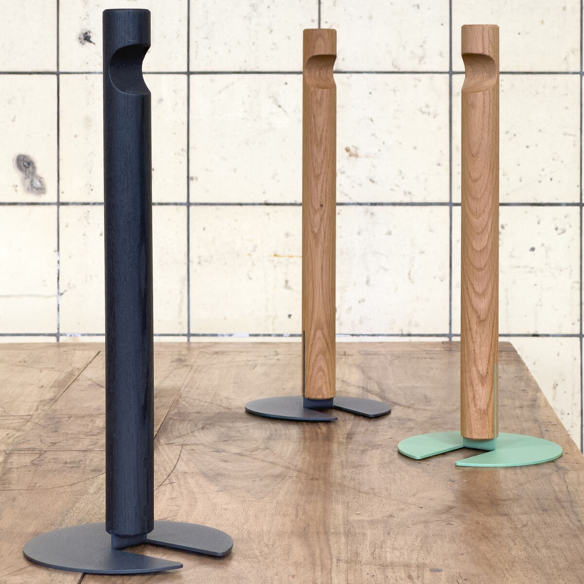 Drei Küchenrollenhalter »GIVE« der Marke finbeaux von Christoph Schoen stehen auf einem Holztisch vor einer gefliesten Wand. Zwei Halter sind naturfarben, einer schwarz, die Standfüße sind in Schwarz, Grau und Grün gehalten. Alle zeigen ein minimalistisches, klares Design.