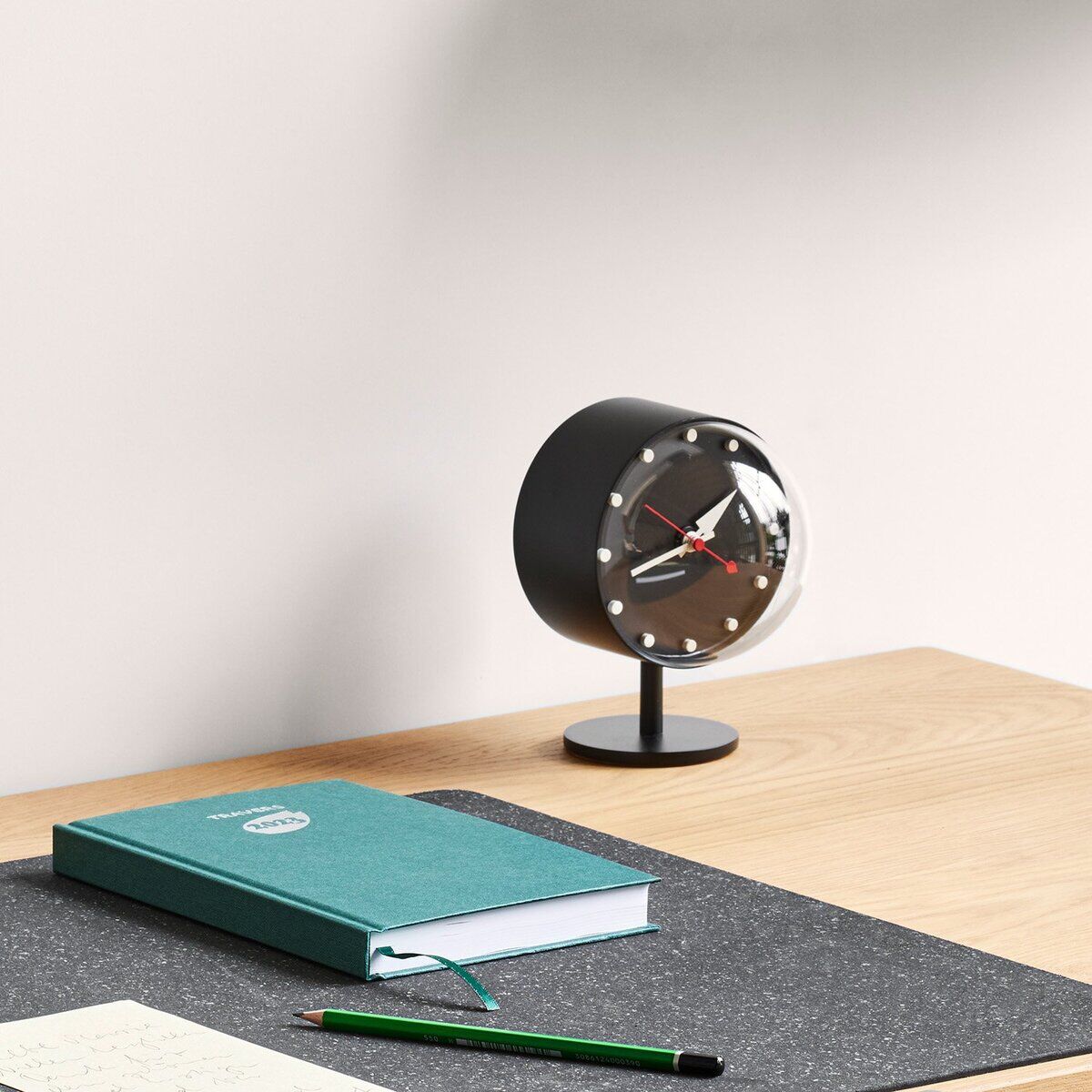 Vitra Tischuhr »Night Clock« von George Nelson steht auf hellem Holzschreibtisch mit schwarzer Unterlage. Runde Uhr mit schwarzem Gehäuse, silbernen Punkten, messingfarbenem Standfuß und Zeigern, daneben türkisfarbenes Notizbuch und grüner Bleistift.