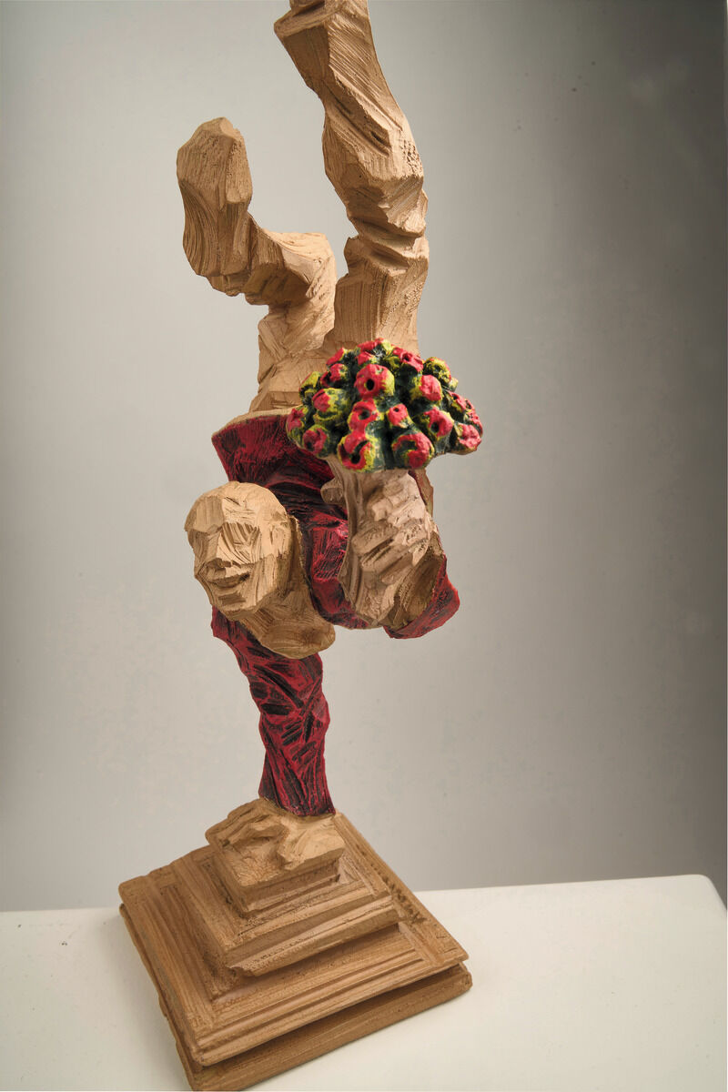 Figurative Skulptur von Roman Johann Strobl in Holzoptik, zeigt eine akrobatische Figur mit Blumenstrauß, auf einem verzierten Sockel stehend. Die Skulptur ist detailliert und in den Farben Holzbraun, Rot und Grün gestaltet.