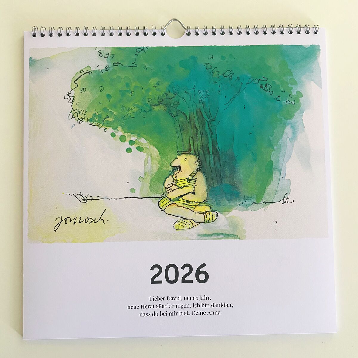 Auf dem Foto ist ein Wandkalender für das Jahr 2026 zu sehen, der im oberen Bereich eine bunte Illustration eines sitzenden, nachdenklichen Wesens unter einem stilisierten Baum zeigt. Darunter steht eine persönliche Widmung und die Zahl 2026 in fetter Schrift. Der Kalender ist an einer hellen Wand aufgehängt.