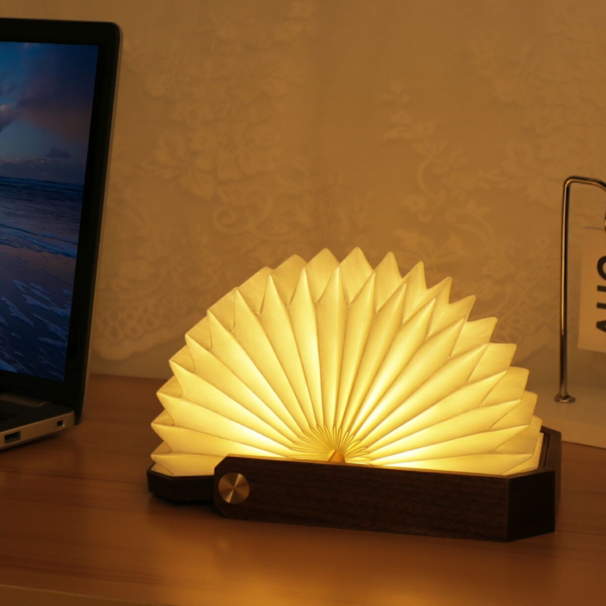 Eine Origami-Lampe aus Holz und Papier steht auf einem Schreibtisch. Sie hat eine gefaltete, ventilatorähnliche Form und strahlt warmes gelbes Licht aus. Im Hintergrund ist ein Laptop mit einem Strandbildschirm und ein Kalendarium mit der Anzeige 'AUG 20' zu sehen.