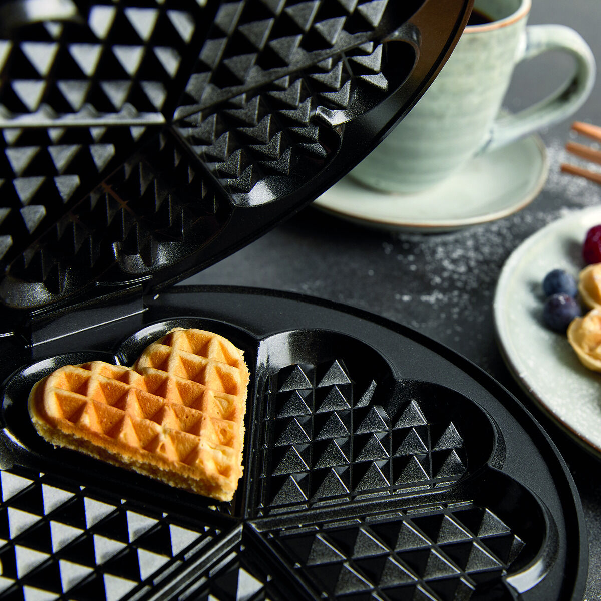 Ein geöffnetes Waffeleisen mit einer herzförmigen Waffel im Inneren. Auf einem Teller daneben liegen weitere goldbraune Herzwaffeln, garniert mit Beeren. Eine Tasse und ein Holzlöffel sind im Hintergrund zu sehen, auf einem dunklen Tisch.