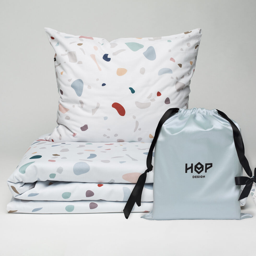 Ein Set aus Bettbezug und Kissenbezug mit Terrazzo-Muster in verschiedenen Pastelltönen auf weißem Hintergrund. Daneben steht ein hellblauer Beutel mit schwarzer Aufschrift 'HOP Design'.