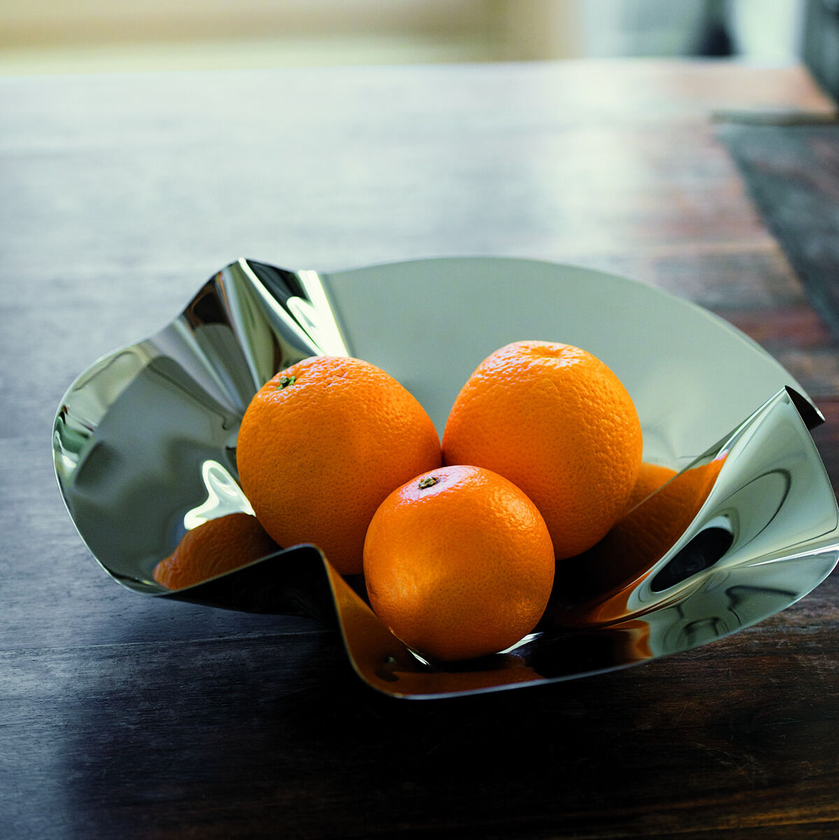 Eine glänzende Metallschale mit dem Namen 'Margarethe', die drei Orangen enthält und auf einem dunklen Holztisch steht, im unscharfen Hintergrund ein heller Raum.