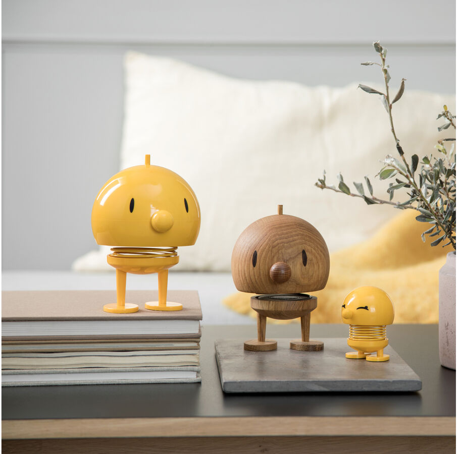 Bild von drei Hoptimist Bumble »Eiche« Figuren, die in einem modernen Wohnzimmer auf Büchern und Steinplatten platziert sind. Die mittlere Figur ist aus hellem Holz gefertigt, während die anderen beiden in verschiedenen Gelbtönen leuchten.