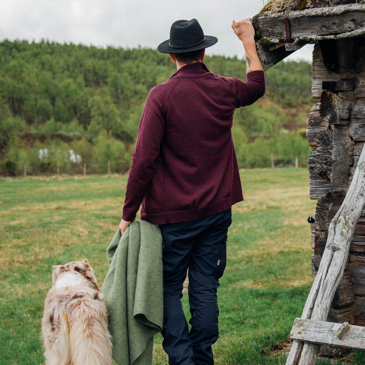 Ein Mann in einem burgunderfarbenen Pullover und schwarzem Hut steht neben einem alten Holzgebäude auf einer grünen Wiese und hält eine grüne Decke in der Hand. Ein Hund steht neben ihm. Im Hintergrund gibt es einen bewaldeten Hügel unter einem wolkigen Himmel.