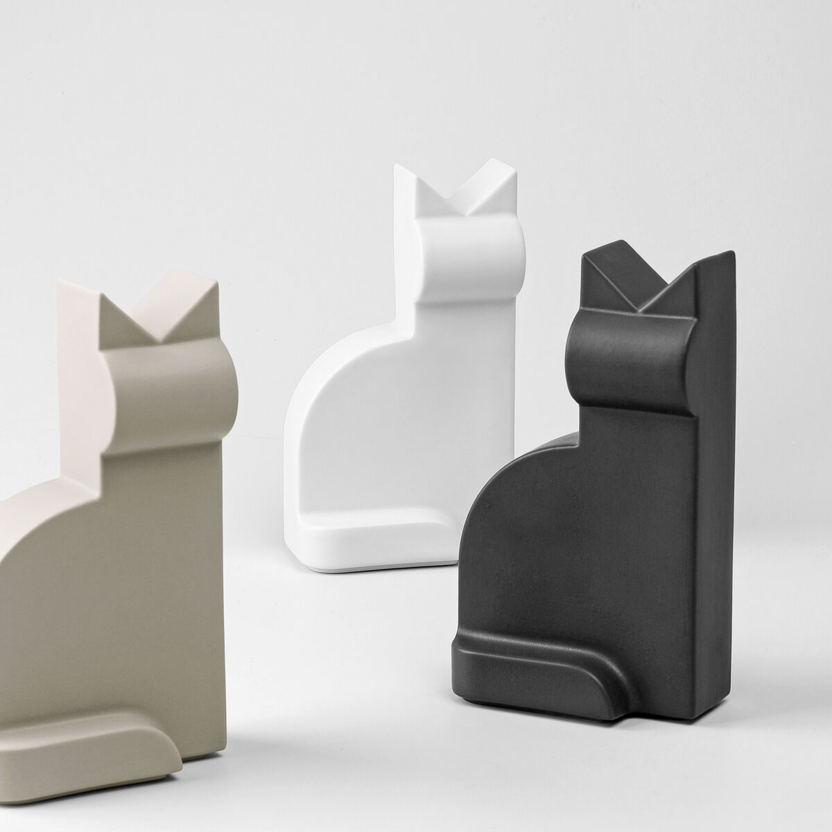 Drei minimalistische Porzellanfiguren in Form von Katzen in den Farben Weiß, Schwarz und Beige auf einer weißen Oberfläche.