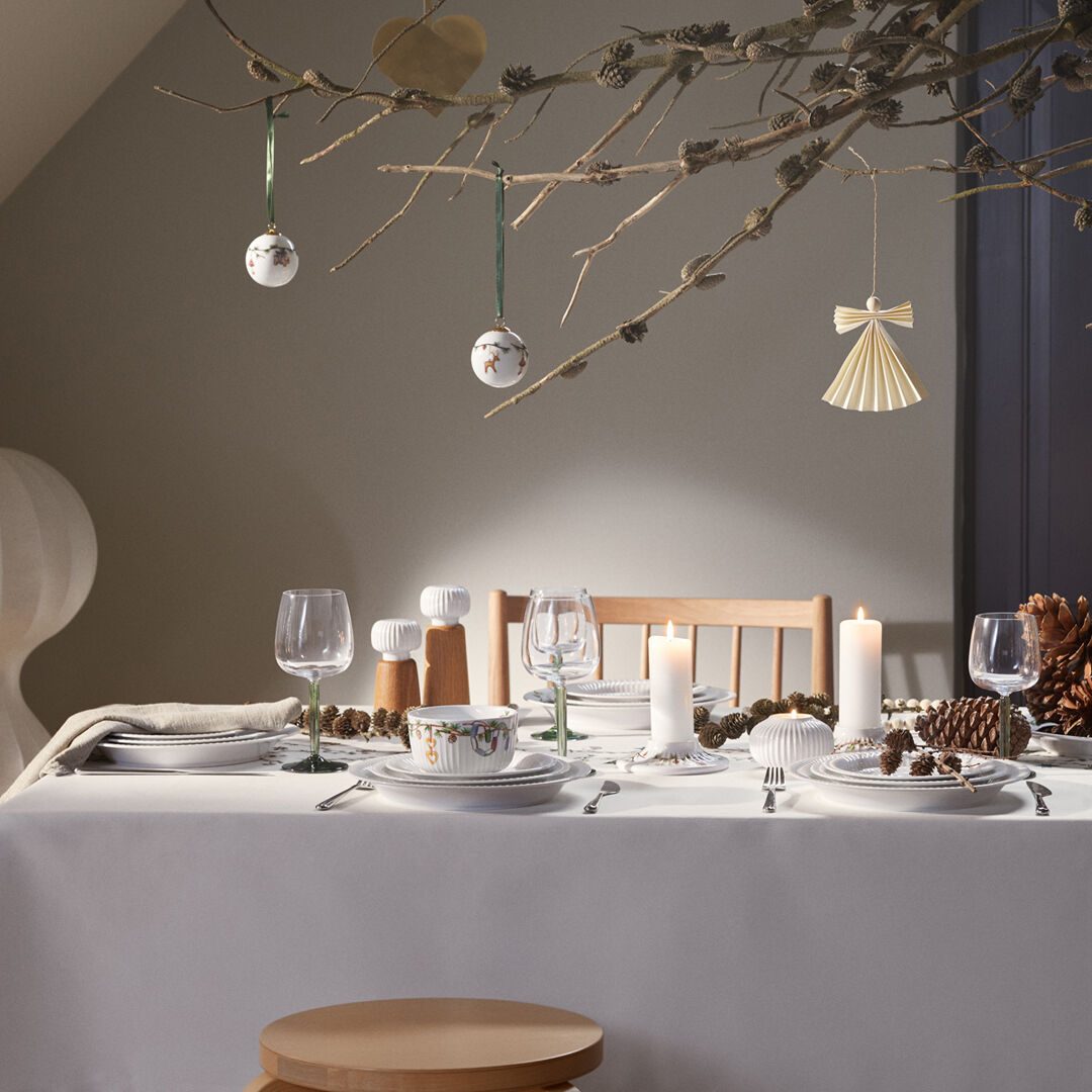 Skandinavisch dekorierter Esstisch mit weißer Tischdecke, Gewürzmühle »Hammershøi« H14, Kerzen, Tannenzapfen und winterlichem Baumschmuck, geeignet für festliche Anlässe wie Weihnachten.