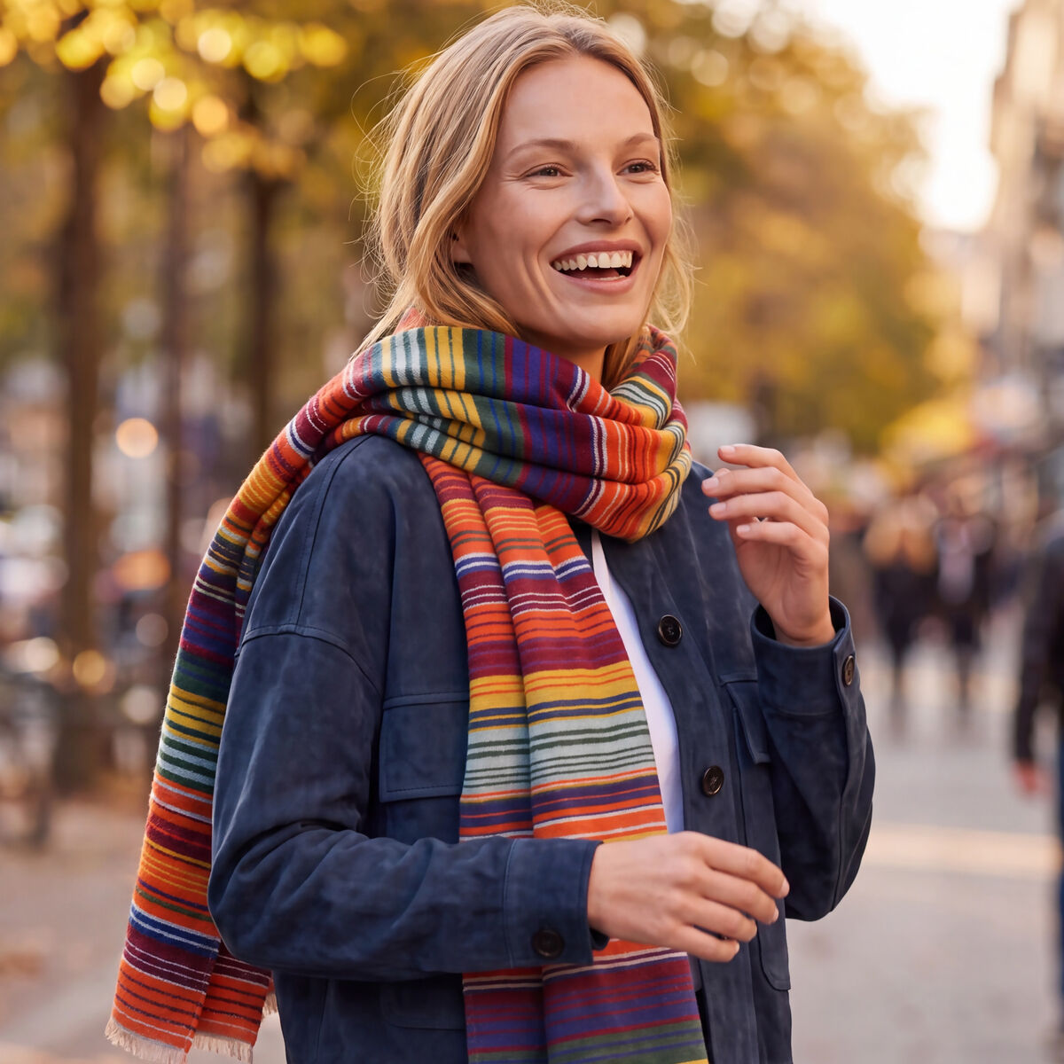 Eine lächelnde Frau steht im Freien auf einer herbstlichen Straße und trägt einen auffällig gestreiften Wollschal »Ariana« von kräftigen Farben wie Rot, Orange, Gelb und Grün. Dazu kombiniert sie eine dunkelblaue Jacke, ein weißes Shirt und blaue Jeans. Die Sonne scheint durch die Bäume.