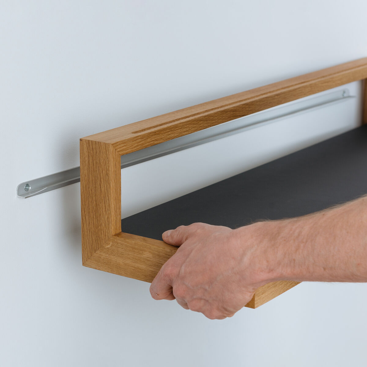 Das Ablagebord »buck« Größe 1 der Marke das kleine b wird mit verdeckter Aluminiumleiste an einer weißen Wand montiert. Die Ablage besteht aus geöltem Eichenholz mit anthrazitfarbener Möbellinoleum-Oberfläche, eine Hand richtet das Board aus.