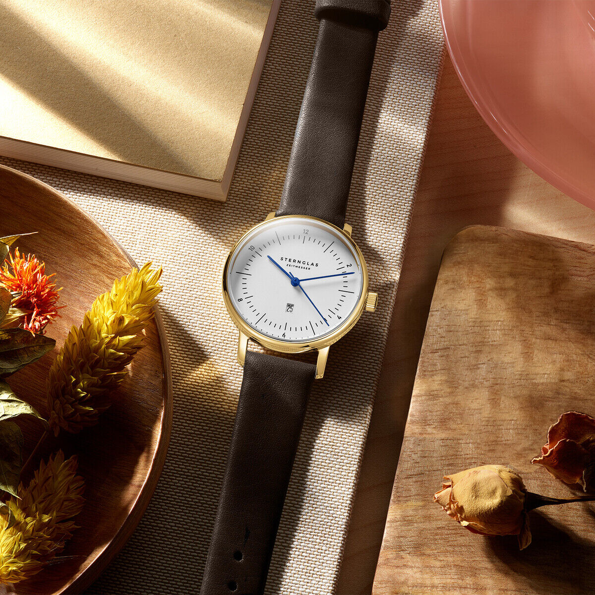 Die Abbildung zeigt eine elegante Armbanduhr der ZEIT-Sonderedition »Naos Xs« mit weißem Zifferblatt, goldfarbenem Gehäuse und schwarzem Lederarmband. Die Uhr liegt dekorativ auf einem Holztablett mit Trockenblumen und weiteren dekorativen Elementen im Hintergrund.