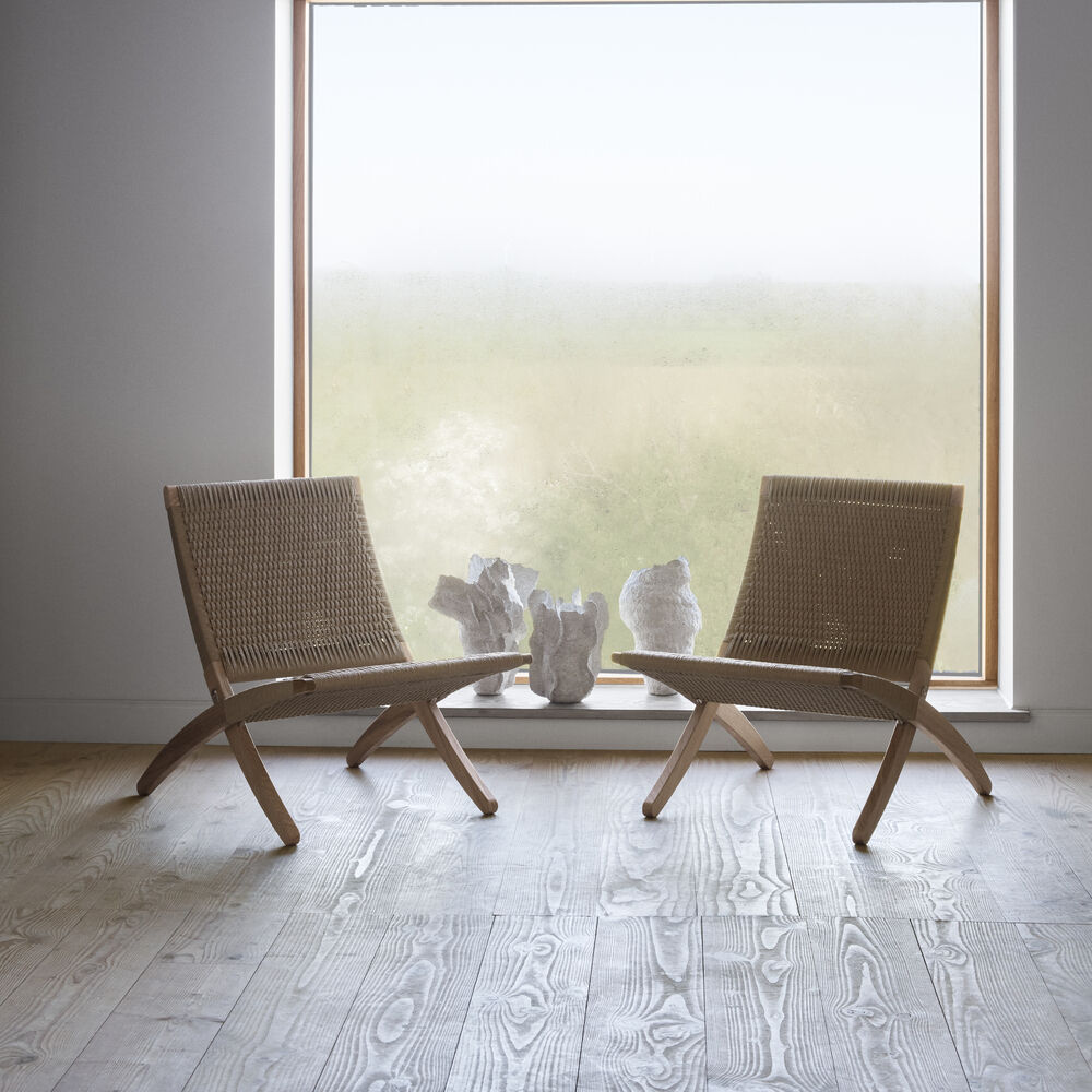 Zwei MG501 Cuba Chairs von Carl Hansen & Søn aus geölter Eiche und hellem Papiergarn-Geflecht stehen auf hellem Holzboden vor großem, rahmenlosem Fenster. Im Hintergrund dekorative Steinskulpturen auf der Fensterbank, natürliches Tageslicht erhellt den Raum.