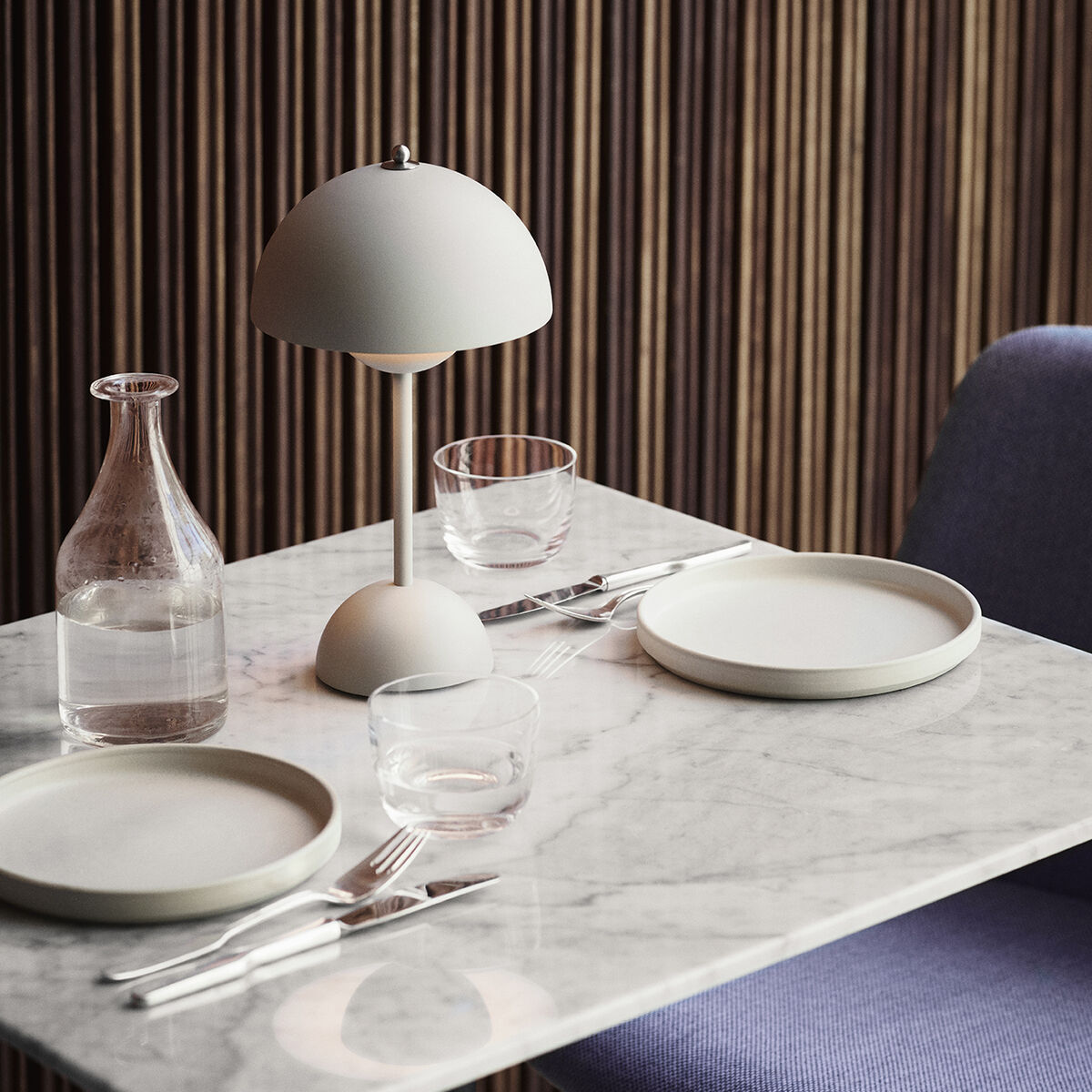 Eine moderne Tischleuchte mit Akku, die auf einem Esstisch mit Marmordekor steht. Die Leuchte hat einen runden weißen Schirm und ein minimalistisches Design. Im Hintergrund befinden sich eine graue Wand und ein gepolsterter Stuhl. Auf dem Tisch stehen zwei Teller, zwei Gläser, eine Glasflasche und Besteck.