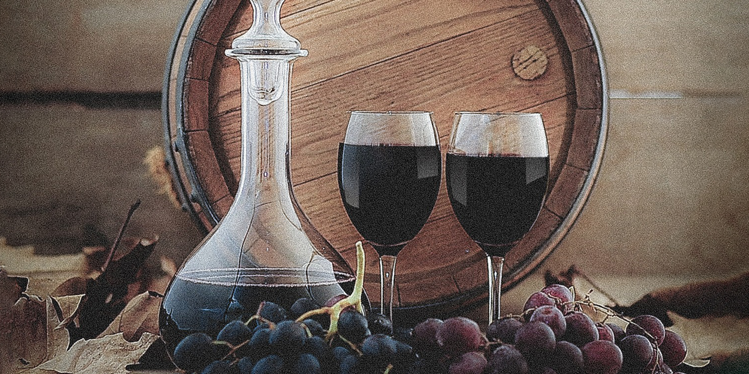 Ein Dekanter aus Glas mit Rotwein, zwei Weingläser mit Rotwein, Trauben und ein Holzfass im Hintergrund.